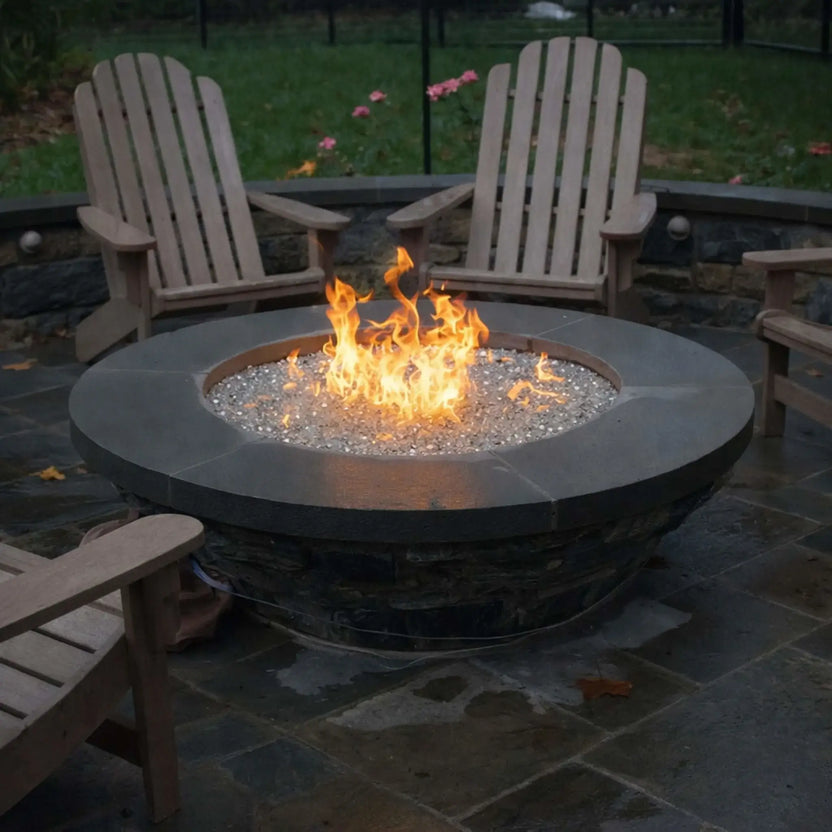 Modern circular fire pit with a smooth dark stone top and clear fire glass, surrounded by wooden Adirondack chairs on a wet patio surface. The flames rise steadily in the center, offering a cozy gathering space in a landscaped backyard with greenery and soft lighting.