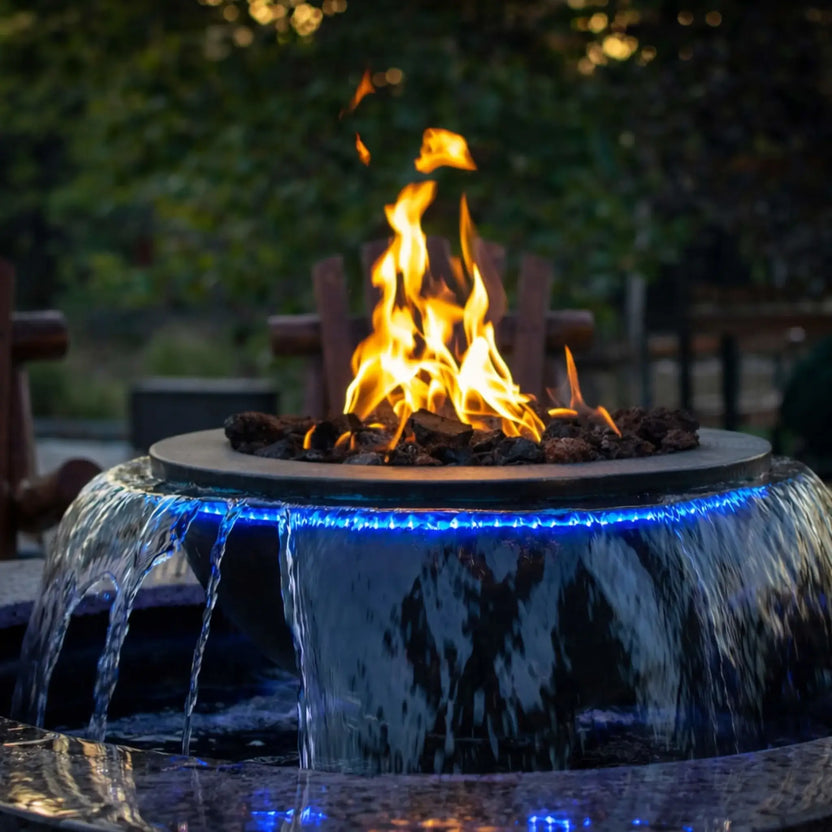 Unique fire and water feature combining a circular fire bowl with cascading water spilling over the edges, illuminated by blue LED lighting. The flame burns above lava rock, creating a striking contrast of fire and water in an upscale outdoor setting.