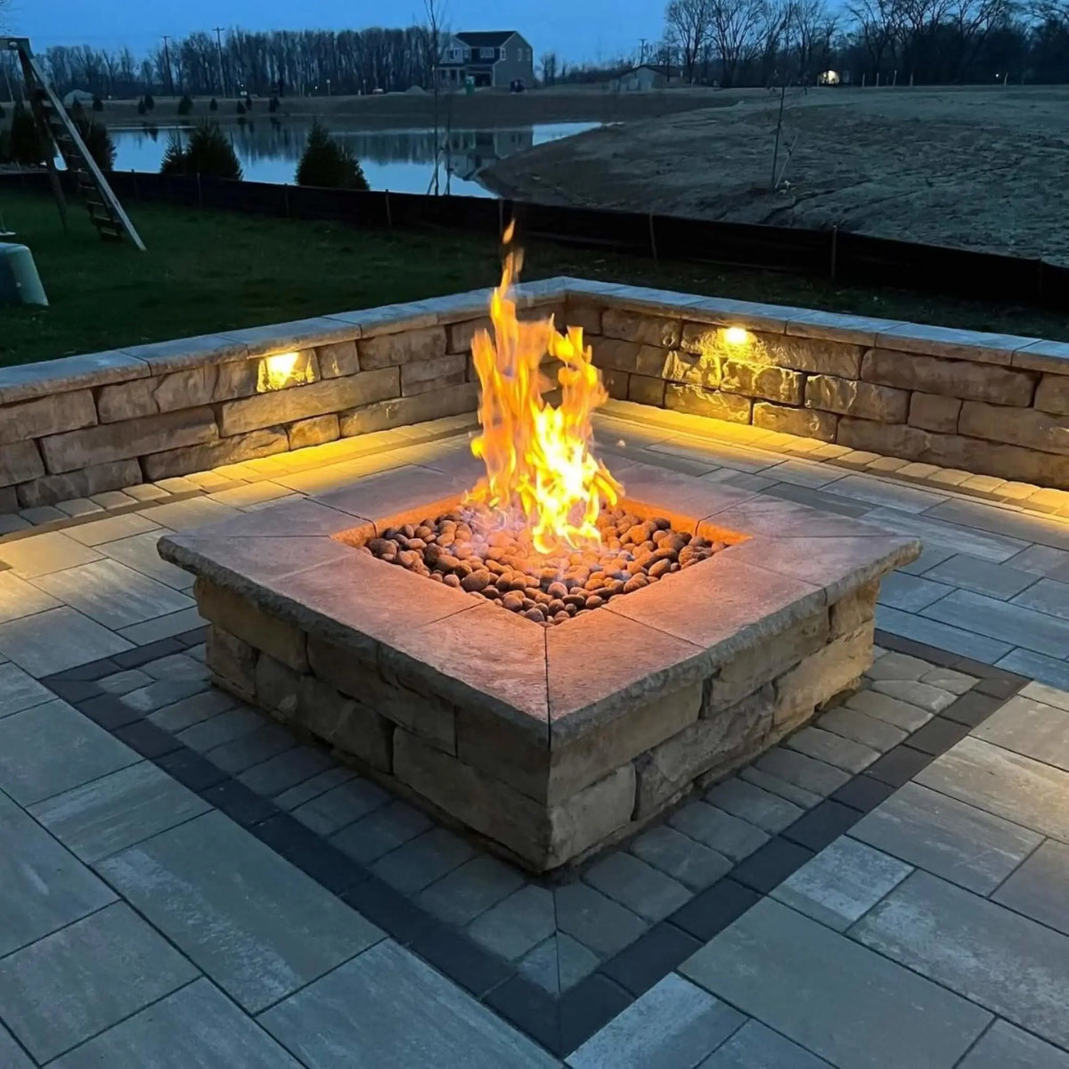 A square stone fire pit powered by the HPC Fire Hearth Products Controls Square Fire Pit Burner Flat Pan FPPK Push Button Flame Sensing FLEX system, glowing warmly at dusk. Flames rise over natural lava rock, surrounded by a paver patio and illuminated seating wall, overlooking a peaceful backyard pond for a relaxed outdoor atmosphere.