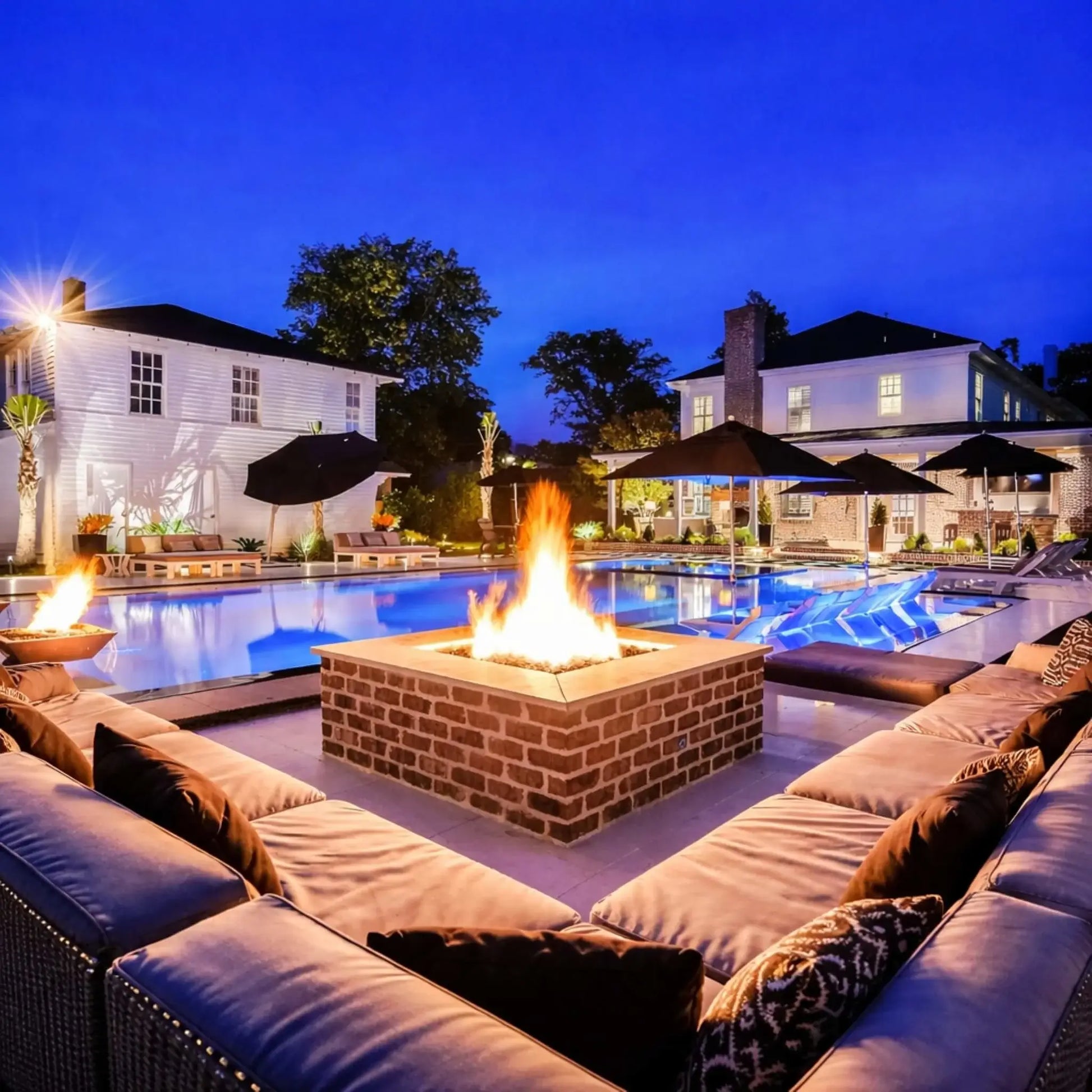 A luxury outdoor poolside scene with a square brick fire pit equipped with the HPC Fire Hearth Products Controls Square Fire Pit Burner Flat Pan FPPK Push Button Flame Sensing FLEX system. The fire glows brightly at night, surrounded by plush seating, umbrellas, and a lit swimming pool, creating a high-end entertaining space.
