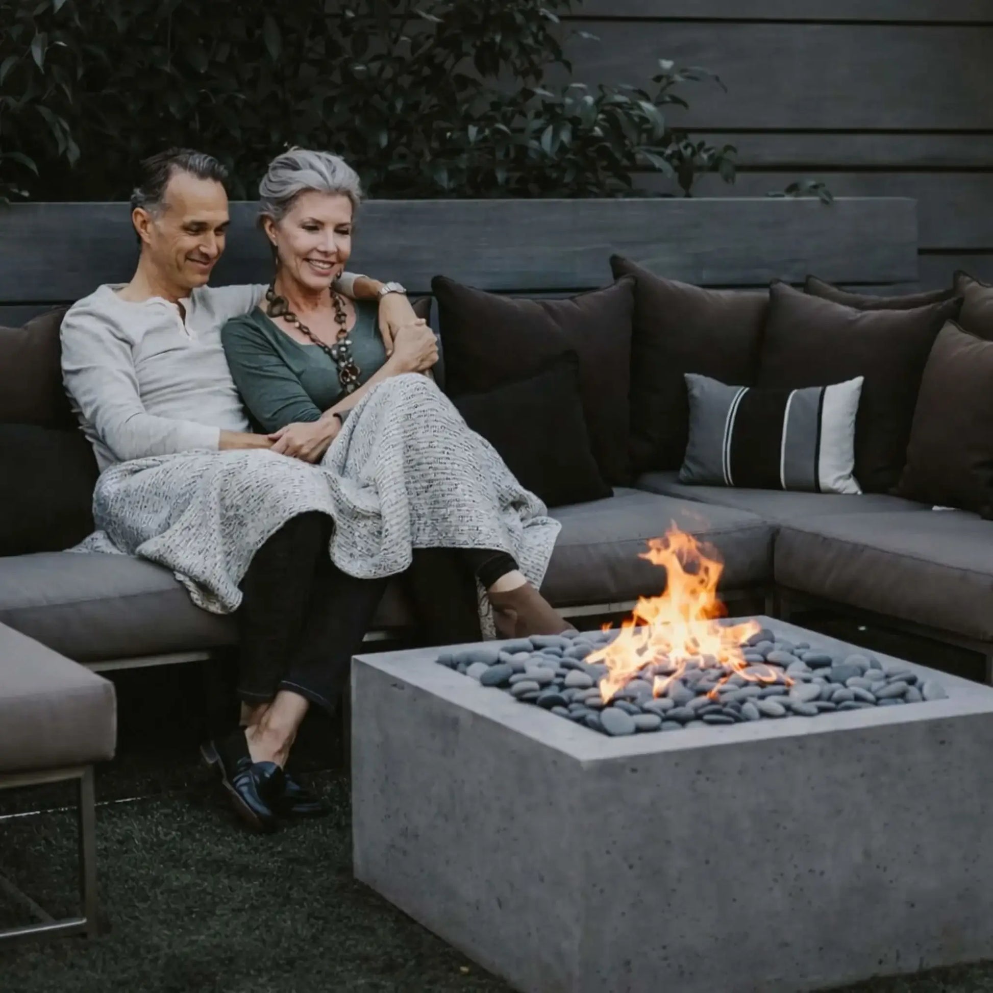 A modern concrete fire pit table showcasing the HPC Fire Hearth Products Controls Square Fire Pit Burner Flat Pan FPPK Push Button Flame Sensing FLEX burner. A couple relaxes on a sectional sofa, enjoying flames over smooth gray stones, creating a cozy and intimate outdoor seating area with a contemporary feel.