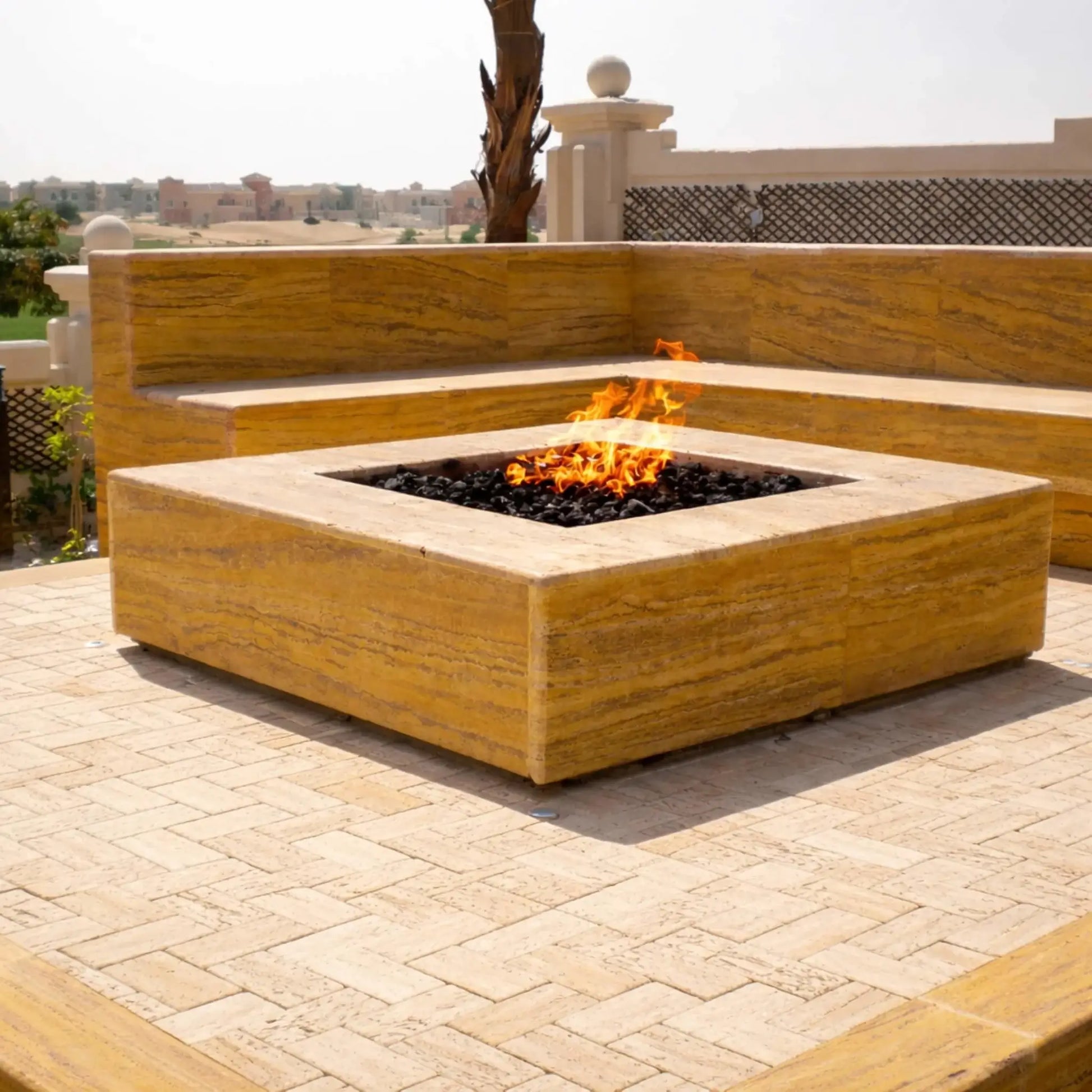 A sleek, modern square fire pit constructed with smooth tan stone, showcasing the HPC Fire Hearth Products Controls Square Fire Pit Burner Flat Pan Match Lit CSA MLFPK FLEX burner. Black fire glass enhances the flame contrast, set within a contemporary outdoor lounge space with built-in bench seating and desert-style surroundings.
