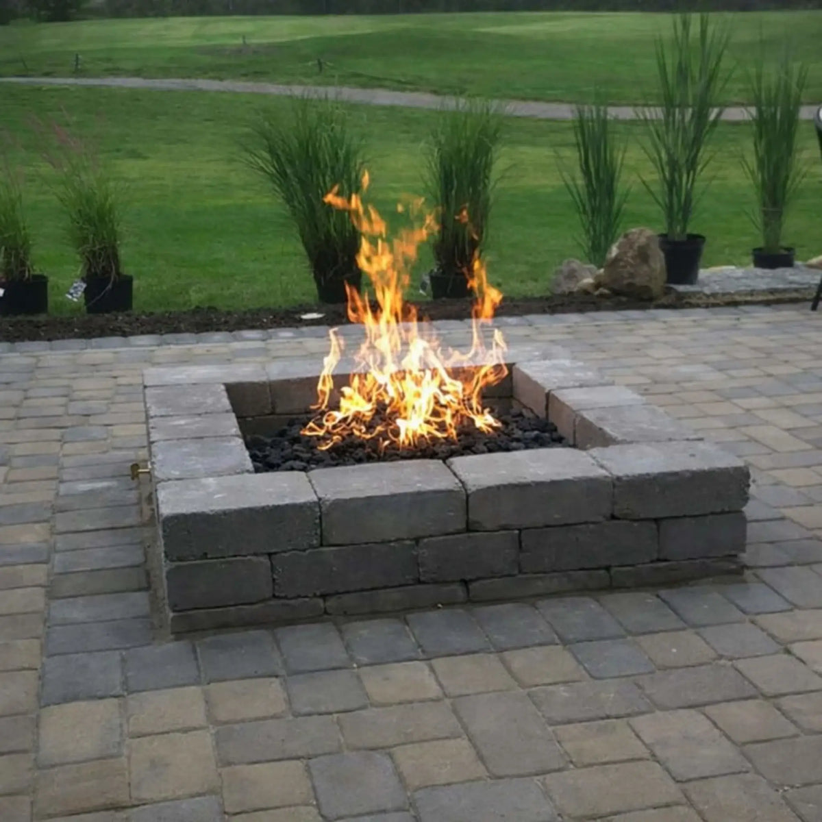 Classic backyard paver patio with a square block fire pit burning bright flames over dark fire media, demonstrating the HPC Fire Hearth Products Controls Square Fire Pit Burner Flat Pan Standard Torpedo Penta Burner Electronic Ignition system. The surrounding lawn and decorative plants create a natural residential setting ideal for outdoor gatherings.