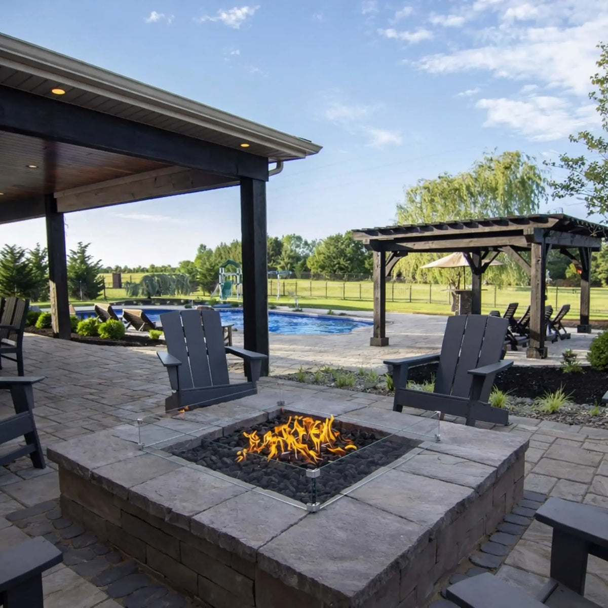 Modern backyard patio featuring a square gas fire pit with black lava rock and a clear tempered glass wind guard, surrounded by gray Adirondack chairs on a stone paver surface. A covered patio structure and pergola sit nearby, with a swimming pool and open green landscape in the background, creating a relaxed outdoor living space ideal for entertaining.