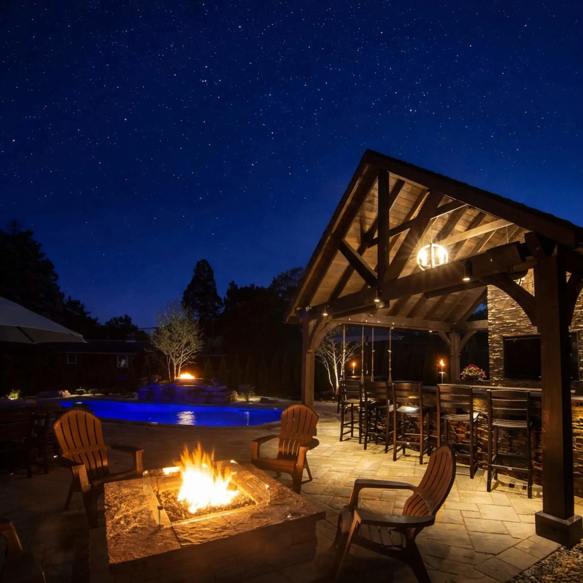 Evening outdoor living area with a glowing square fire pit encased in a tempered glass wind guard, set on a stone patio beside a lit swimming pool. Wooden Adirondack chairs surround the fire, while a large timber-frame pavilion with bar seating and warm lighting creates an inviting nighttime backyard atmosphere under a star-filled sky.