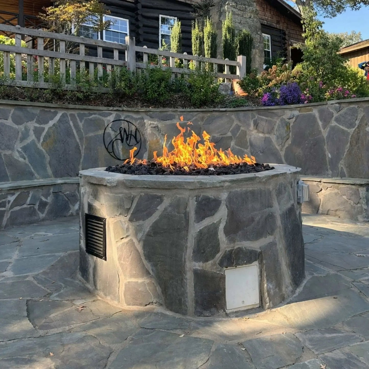 Large custom stone fire pit with a built-in Penta burner producing tall natural gas flames over black lava rock, set in a backyard patio with stone retaining wall, wood fencing, and landscaped garden, showcasing a residential outdoor fire feature installation.