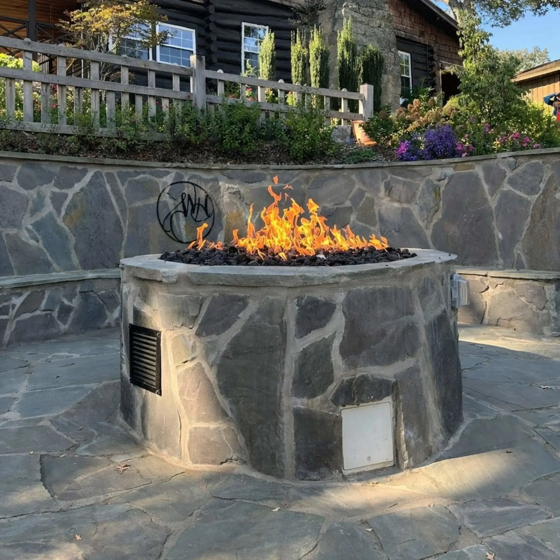 Large custom stone fire pit with a built-in Penta burner producing tall natural gas flames over black lava rock, set in a backyard patio with stone retaining wall, wood fencing, and landscaped garden, showcasing a residential outdoor fire feature installation.