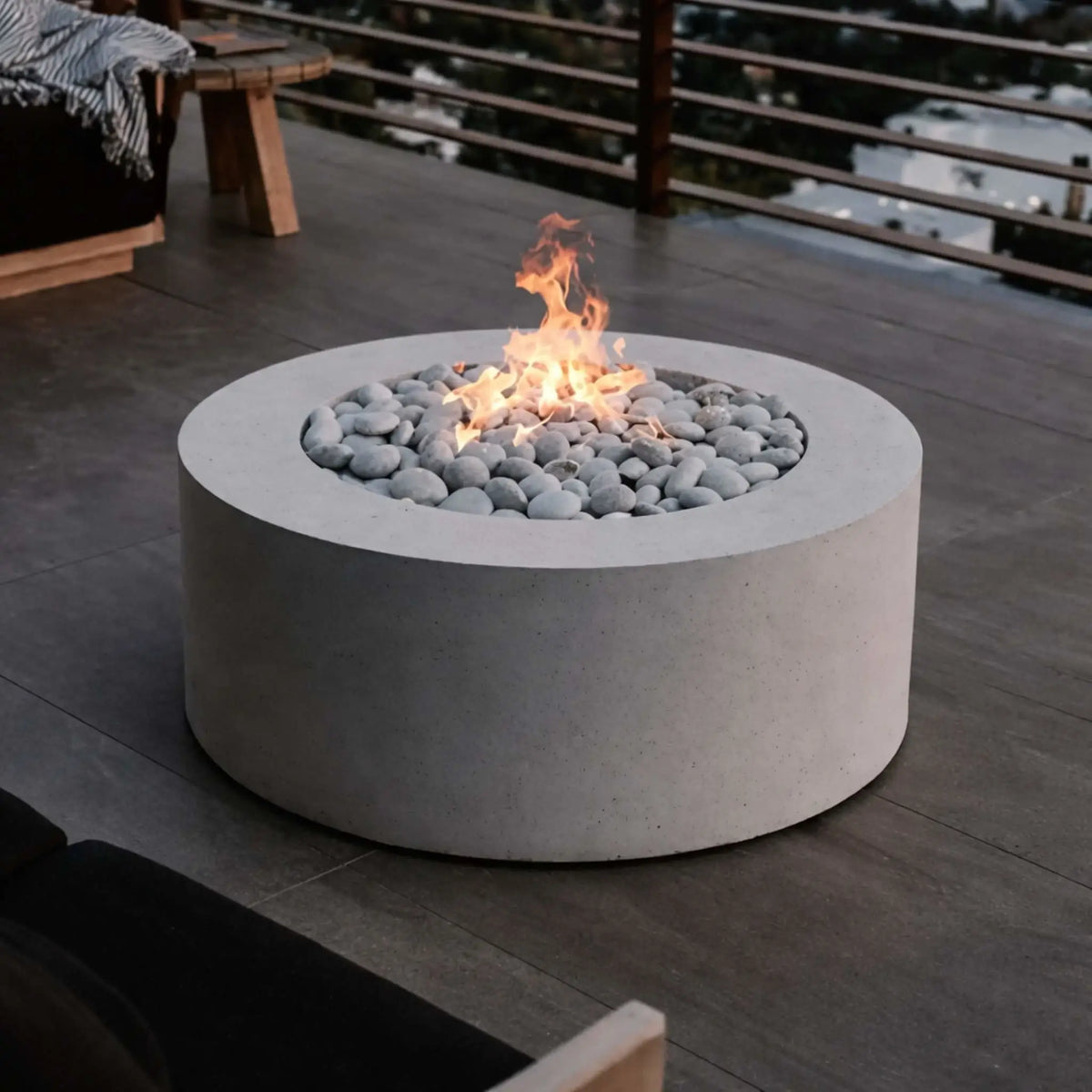 Modern round concrete fire pit table filled with smooth gray river stones and a natural gas flame, set on a sleek rooftop deck with wood plank flooring and horizontal cable railing, overlooking a blurred cityscape at dusk for a contemporary outdoor living space.
