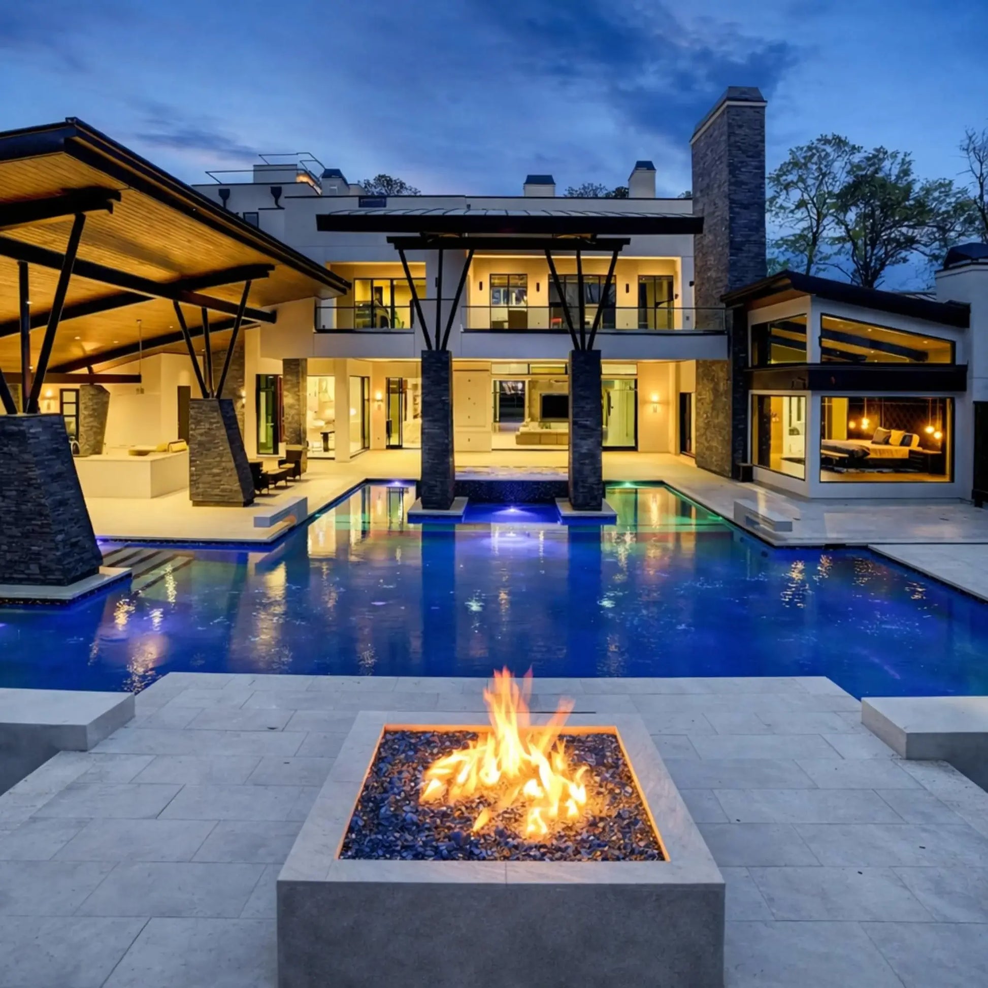Luxury modern home with expansive outdoor living area featuring a rectangular gas fire pit filled with blue fire glass, positioned beside a large illuminated swimming pool, covered patio, and open-concept architecture during evening hours.