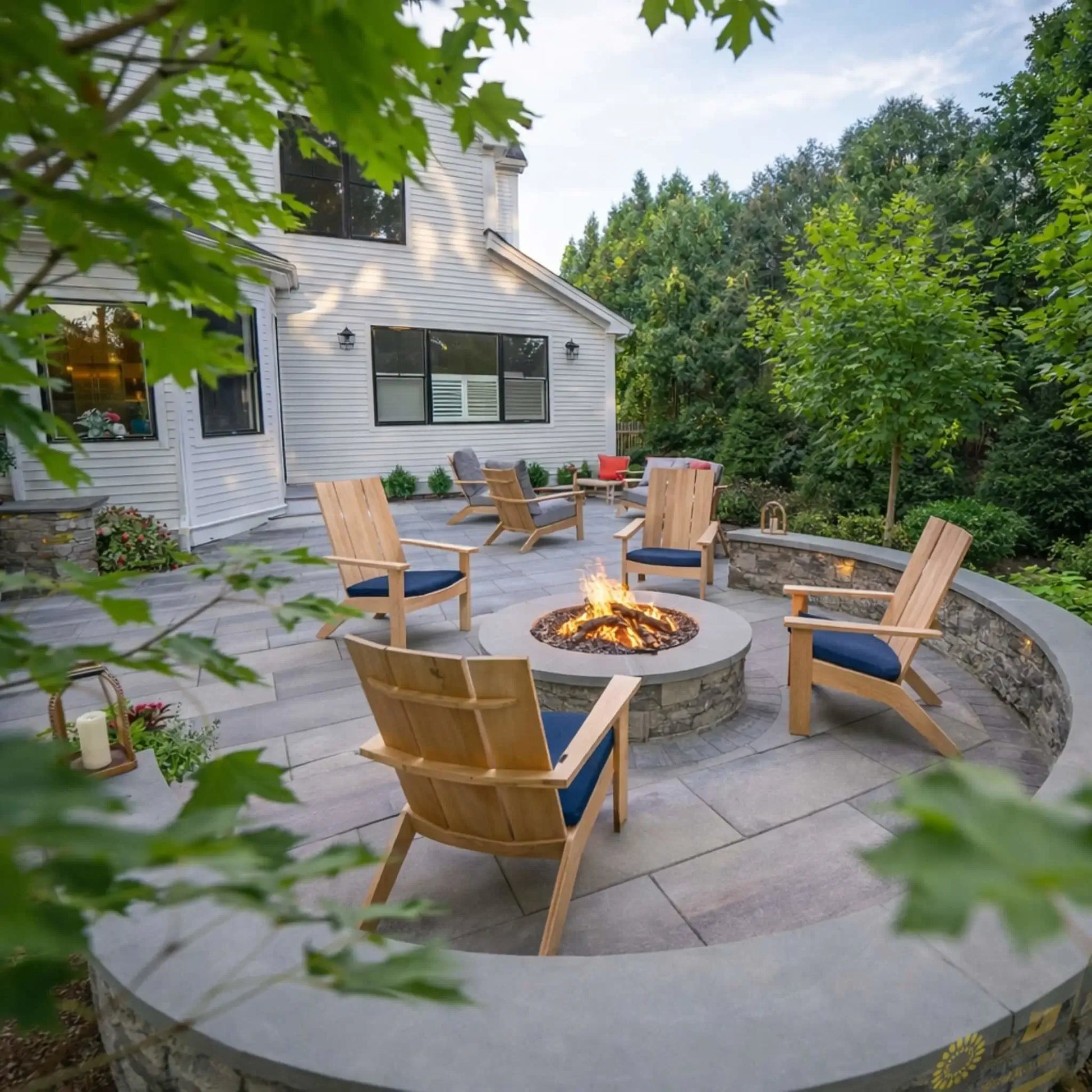 Backyard patio with a circular stone fire pit surrounded by wooden Adirondack chairs, built into a curved seating wall with landscaping and mature trees, creating a welcoming outdoor gathering space in a residential setting.
