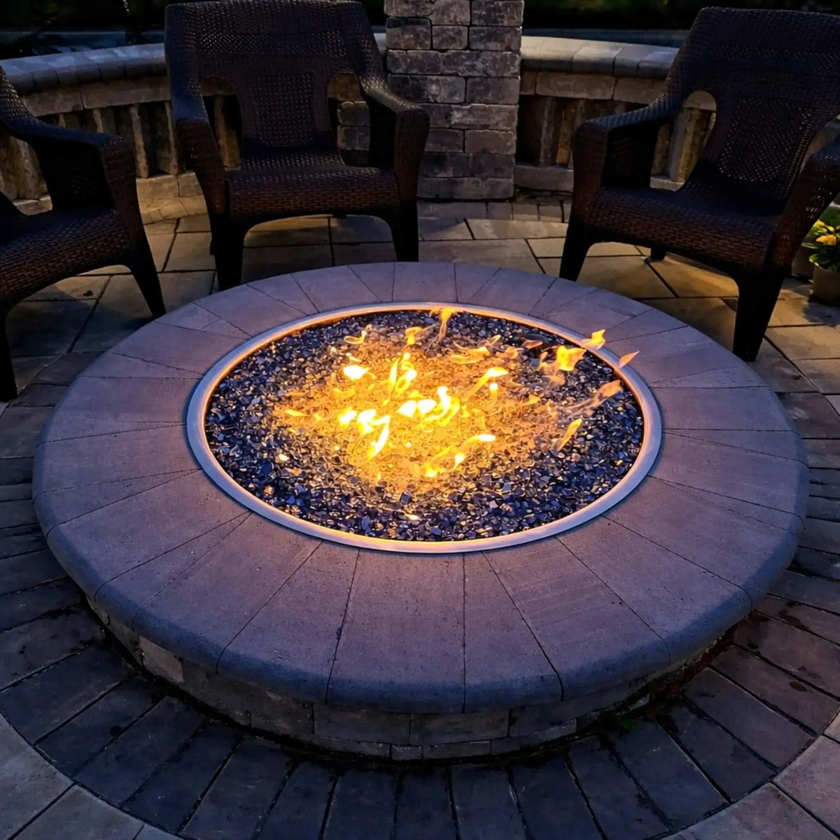 Close-up of a round fire pit with a metal burner ring producing bright flames over reflective fire glass, encircled by woven outdoor chairs on a paved patio, highlighting even flame distribution and modern fire feature design at night.