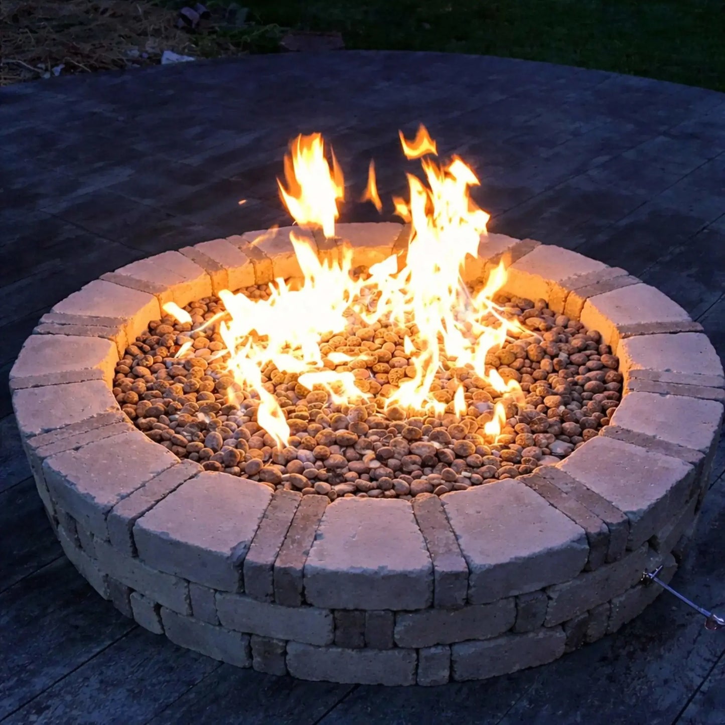 DIY-style circular brick fire pit with stacked stone blocks and lava rock media, showcasing strong natural gas flames on a wooden deck at dusk, emphasizing a simple yet functional backyard fire feature installation.