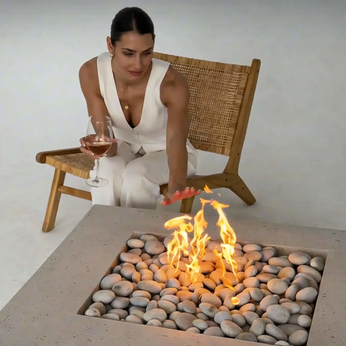Woman seated in a woven lounge chair reaching toward a square concrete fire pit filled with smooth river stones and active flames, holding a glass of wine, illustrating a relaxed indoor or staged outdoor fire feature experience.