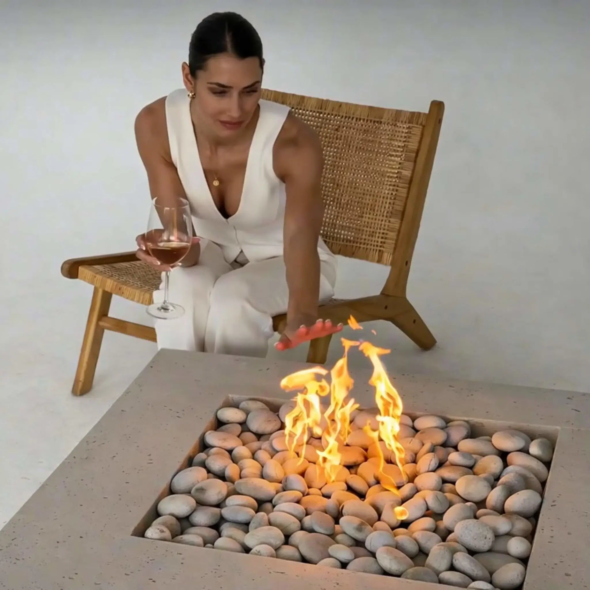 Woman seated in a woven lounge chair reaching toward a square concrete fire pit filled with smooth river stones and active flames, holding a glass of wine, illustrating a relaxed indoor or staged outdoor fire feature experience.