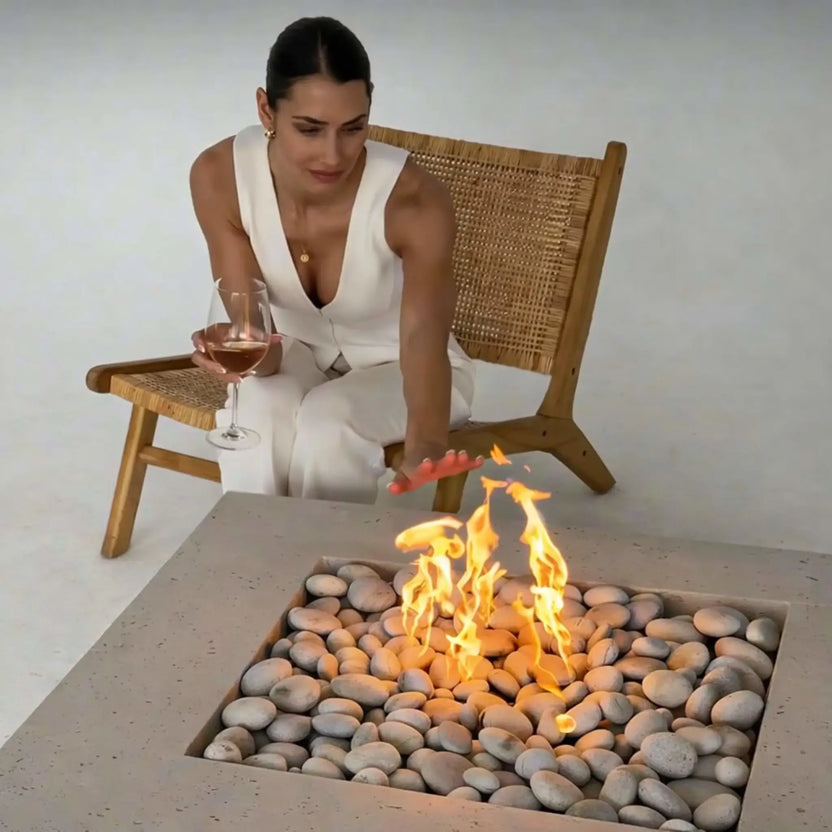 Woman seated in a woven lounge chair reaching toward a square concrete fire pit filled with smooth river stones and active flames, holding a glass of wine, illustrating a relaxed indoor or staged outdoor fire feature experience.