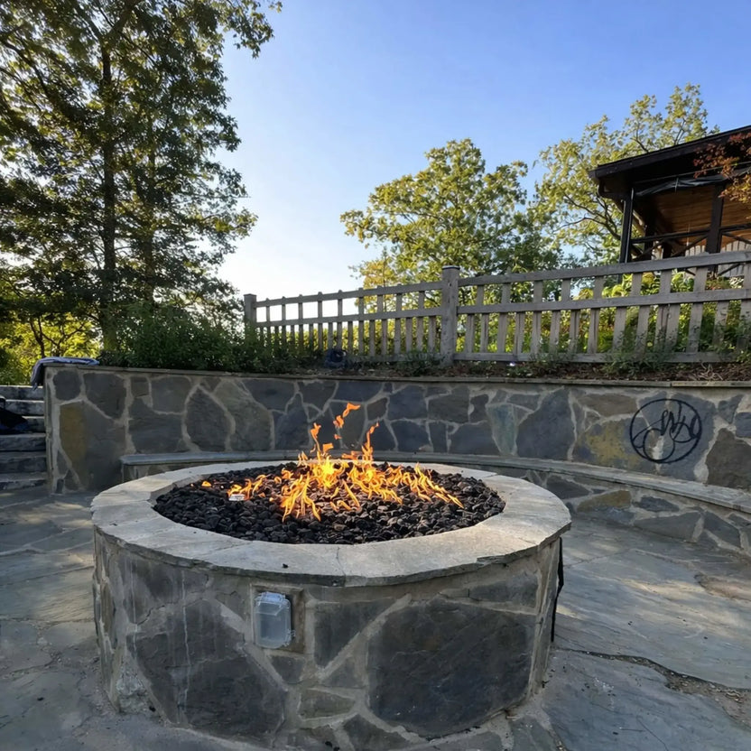 Custom round stone fire pit with black lava rock and wide flame pattern, built into a multi-level patio with stone retaining walls, wood fencing, and surrounding trees, demonstrating a high-end residential outdoor fire installation.