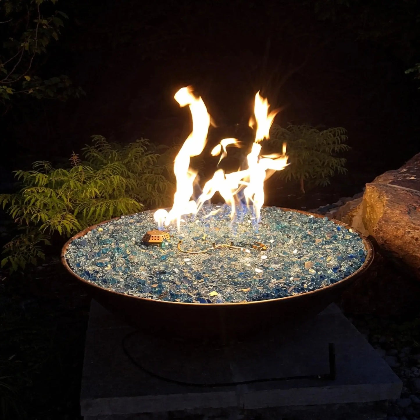 Close-up of a shallow metal fire bowl filled with blue fire glass, showing tall dancing flames against a dark outdoor backdrop with surrounding rocks and foliage, highlighting decorative fire media and burner performance.