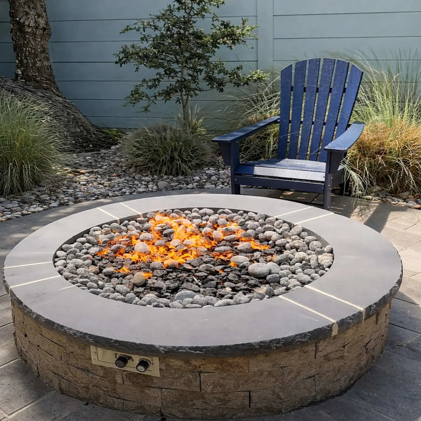 Round stone fire pit with a mix of river rock and lava rock media, set in a landscaped backyard with a blue Adirondack chair, ornamental grasses, and a privacy fence, showcasing a clean and modern residential fire feature.