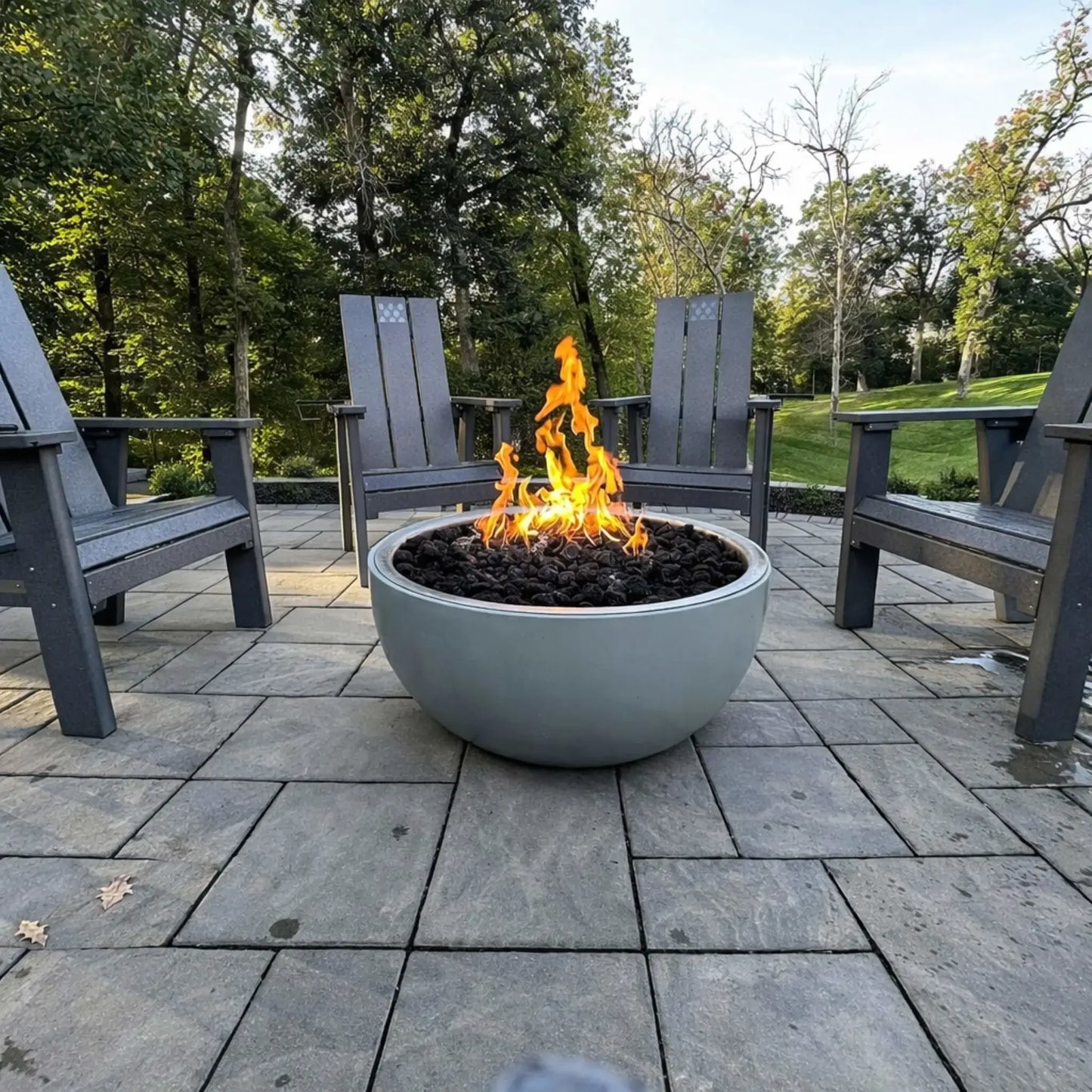 Modern round concrete fire bowl with a Penta-style flame pattern burning over dark fire media, surrounded by gray Adirondack chairs on a paved patio, creating a cozy outdoor seating area in a wooded backyard setting.