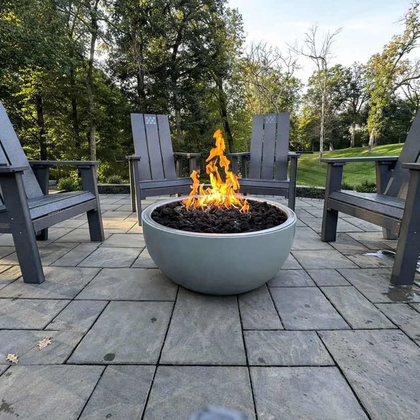Modern round concrete fire bowl with a Penta-style flame pattern burning over dark fire media, surrounded by gray Adirondack chairs on a paved patio, creating a cozy outdoor seating area in a wooded backyard setting.
