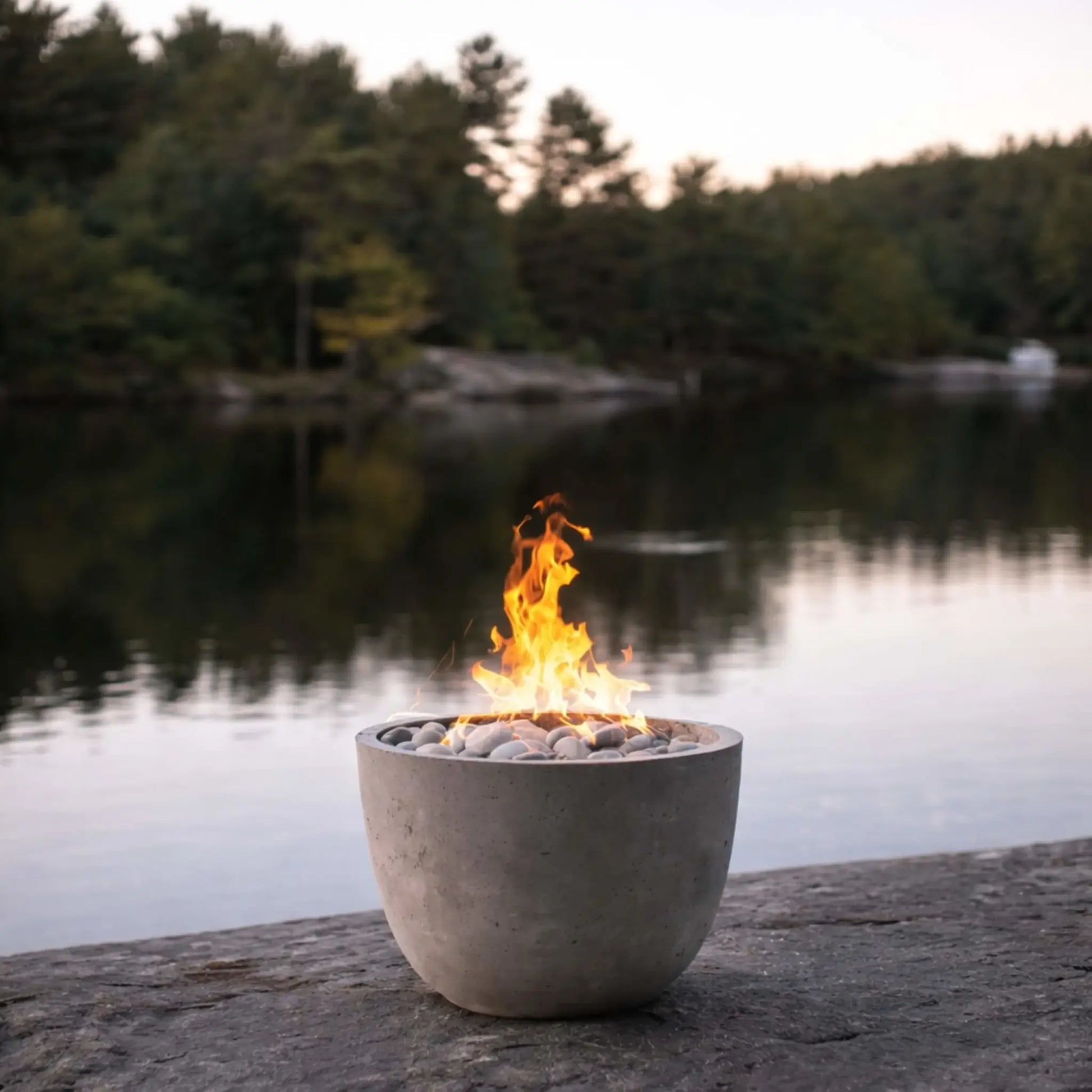 Compact round concrete fire bowl with smooth river rock media and a natural gas flame, placed on a rocky lakeside surface with calm water and dense forest in the background, capturing a peaceful outdoor setting at sunset.
