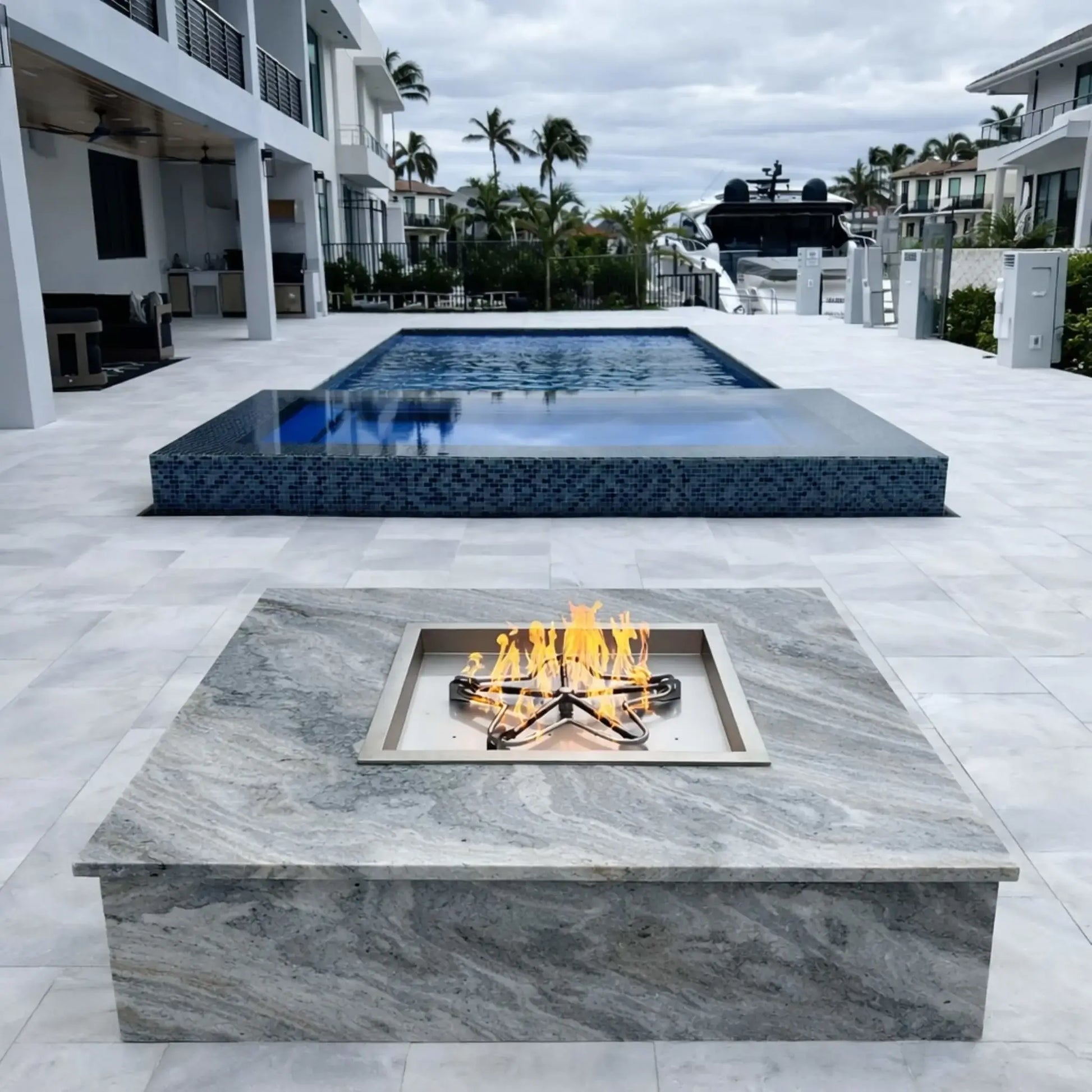 Modern rectangular gas fire pit with a star-shaped burner set into a marble-style surface, positioned in a luxury waterfront backyard with a large swimming pool, palm trees, and contemporary white homes under an overcast sky.