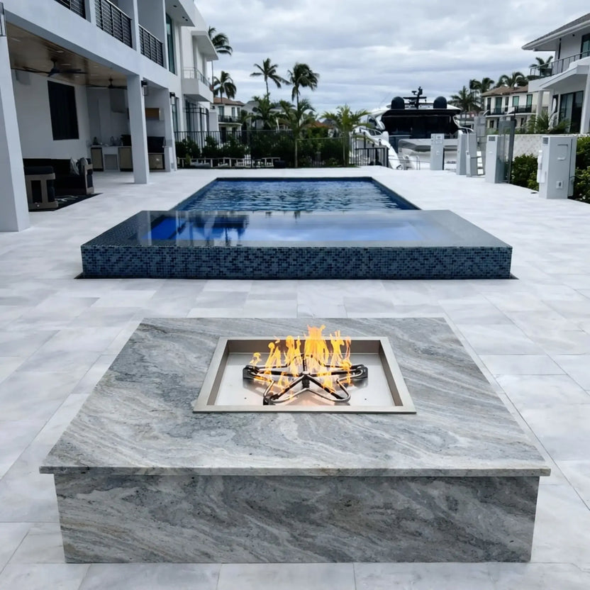 Modern rectangular gas fire pit with a star-shaped burner set into a marble-style surface, positioned in a luxury waterfront backyard with a large swimming pool, palm trees, and contemporary white homes under an overcast sky.