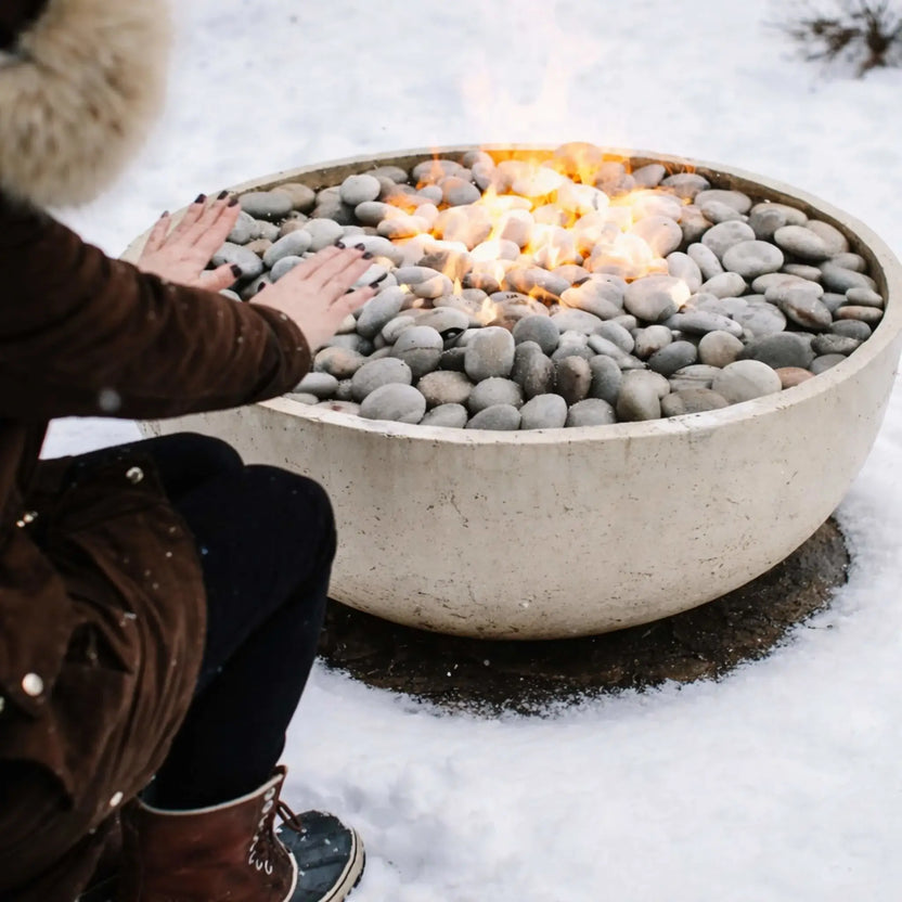 Person warming their hands over a round concrete fire pit filled with smooth gray river stones, surrounded by snow, showcasing active flames and demonstrating cold-weather usability of an outdoor gas fire feature.