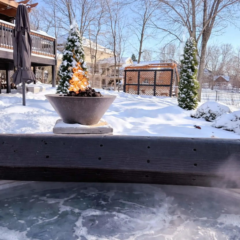 Metal fire bowl with tall flames burning over lava rock, set in a snow-covered backyard near a wooden deck, gazebo structure, and evergreen trees, highlighting a winter-ready outdoor fire feature installation.