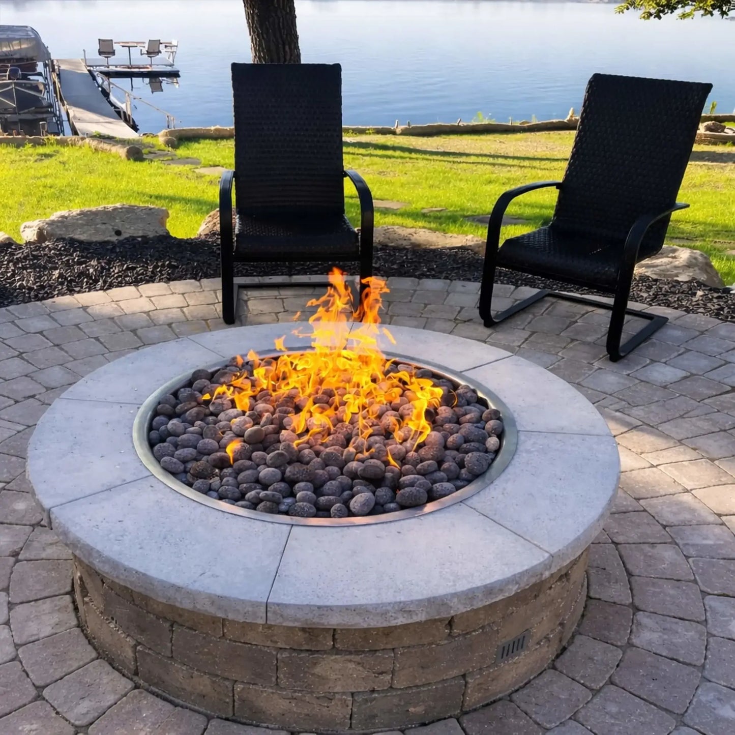 Round stone fire pit with lava rock and bright flames centered on a paver patio, flanked by two black outdoor chairs and overlooking a calm lakeside yard with a dock and lush green lawn.
