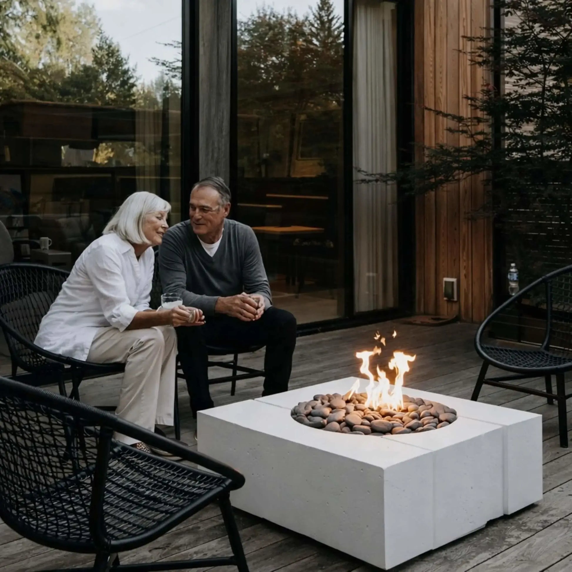 Elderly couple sitting together on a wooden deck beside a square white concrete fire pit filled with river stones and flames, enjoying a relaxed evening in a modern outdoor living space with glass doors and natural wood siding.