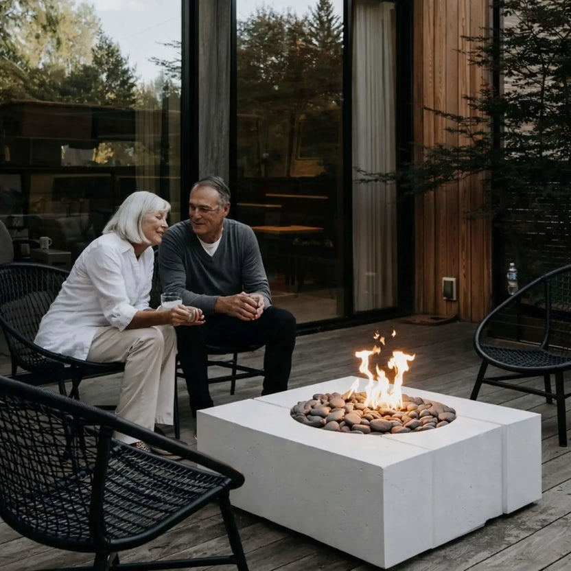 Elderly couple sitting together on a wooden deck beside a square white concrete fire pit filled with river stones and flames, enjoying a relaxed evening in a modern outdoor living space with glass doors and natural wood siding.