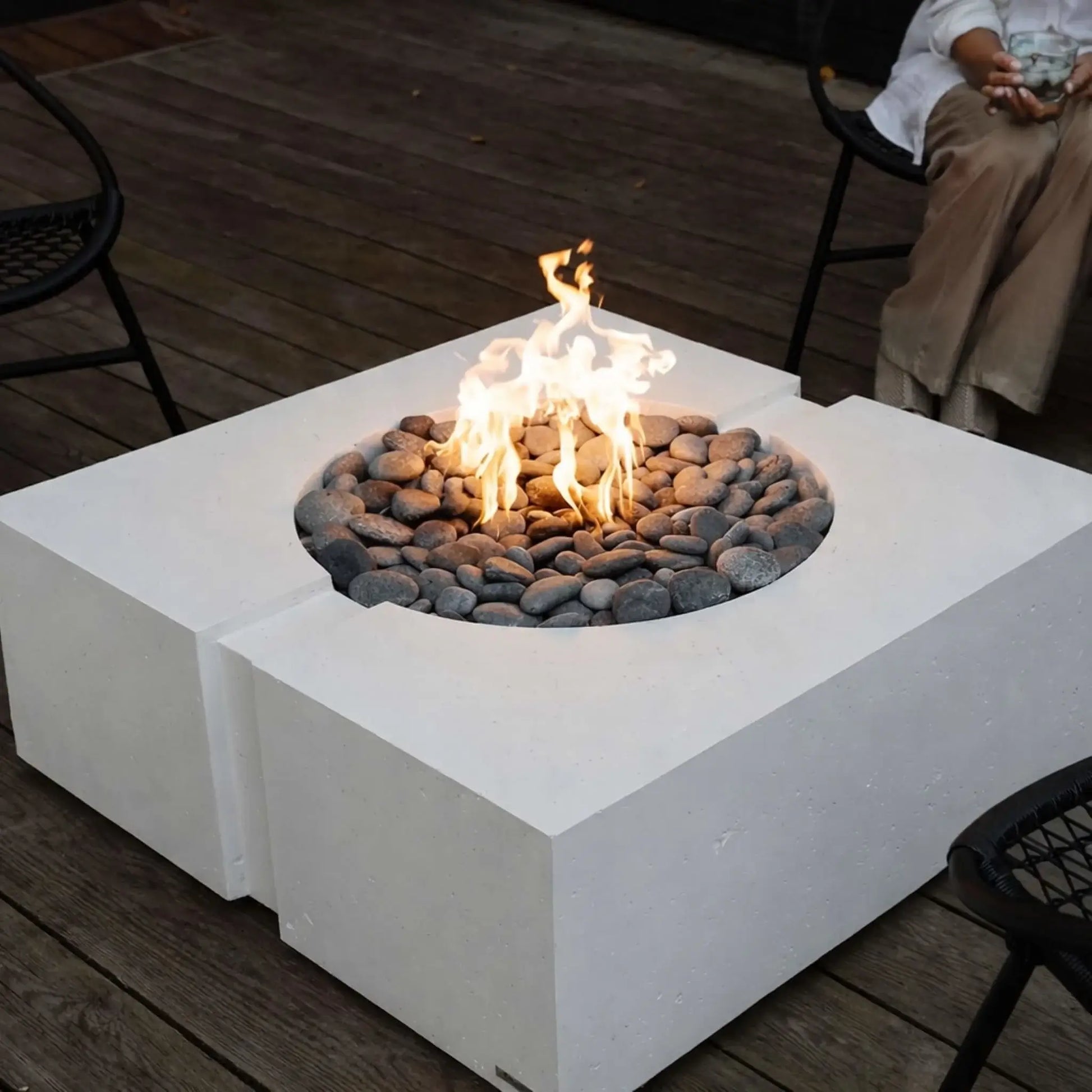 Close-up of a square white concrete fire pit table with a recessed burner and smooth river rock media, showing bright flames on a wooden deck surrounded by black metal patio chairs in a contemporary outdoor setting.