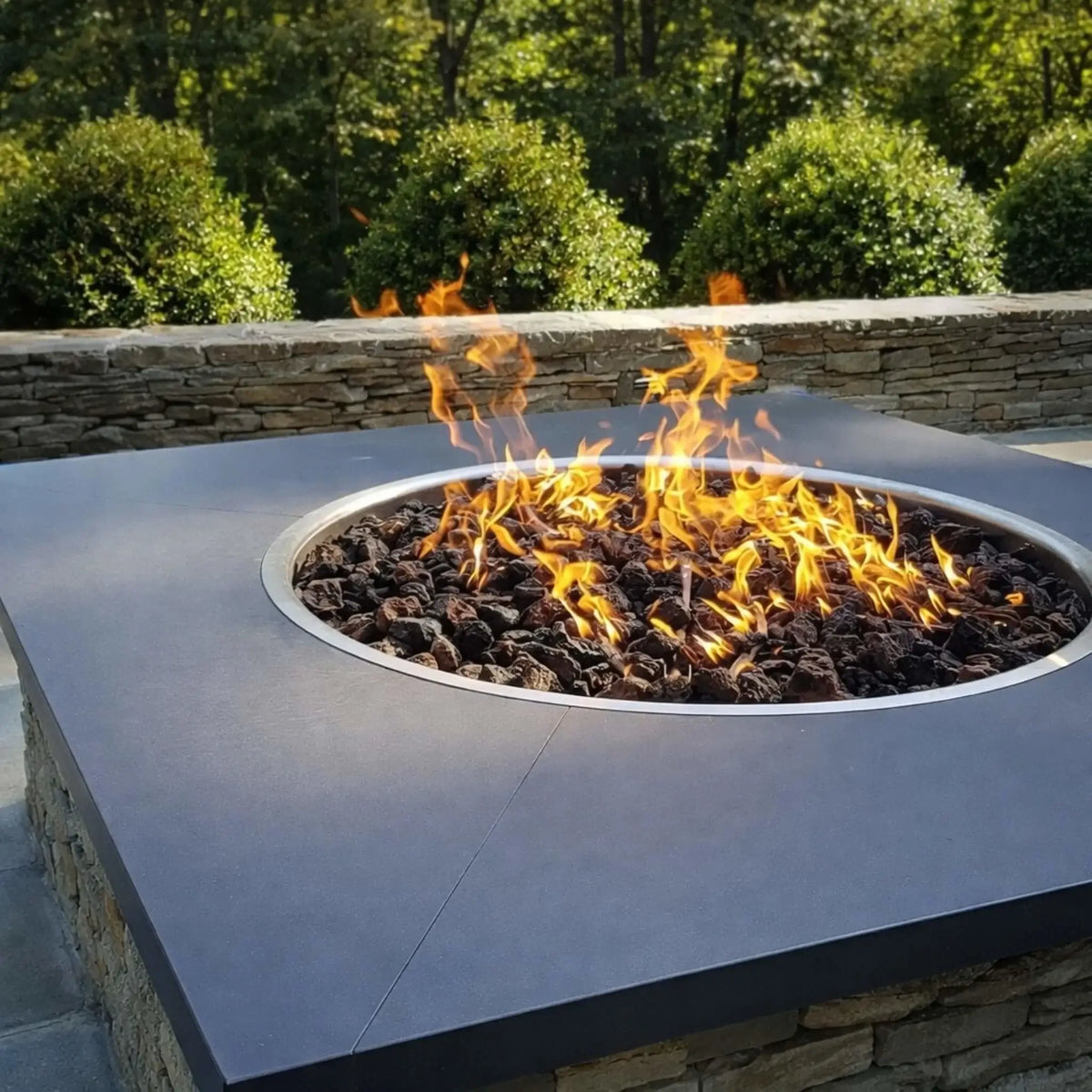 Large square fire pit with a wide metal top and circular burner filled with dark lava rock, producing strong flames in a landscaped backyard with stone retaining wall and neatly trimmed greenery.
