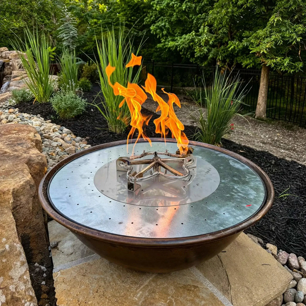 Decorative metal fire bowl with a stainless steel burner plate and visible gas burner assembly, set within a landscaped garden bed featuring ornamental grasses, rocks, and a natural stone water feature.