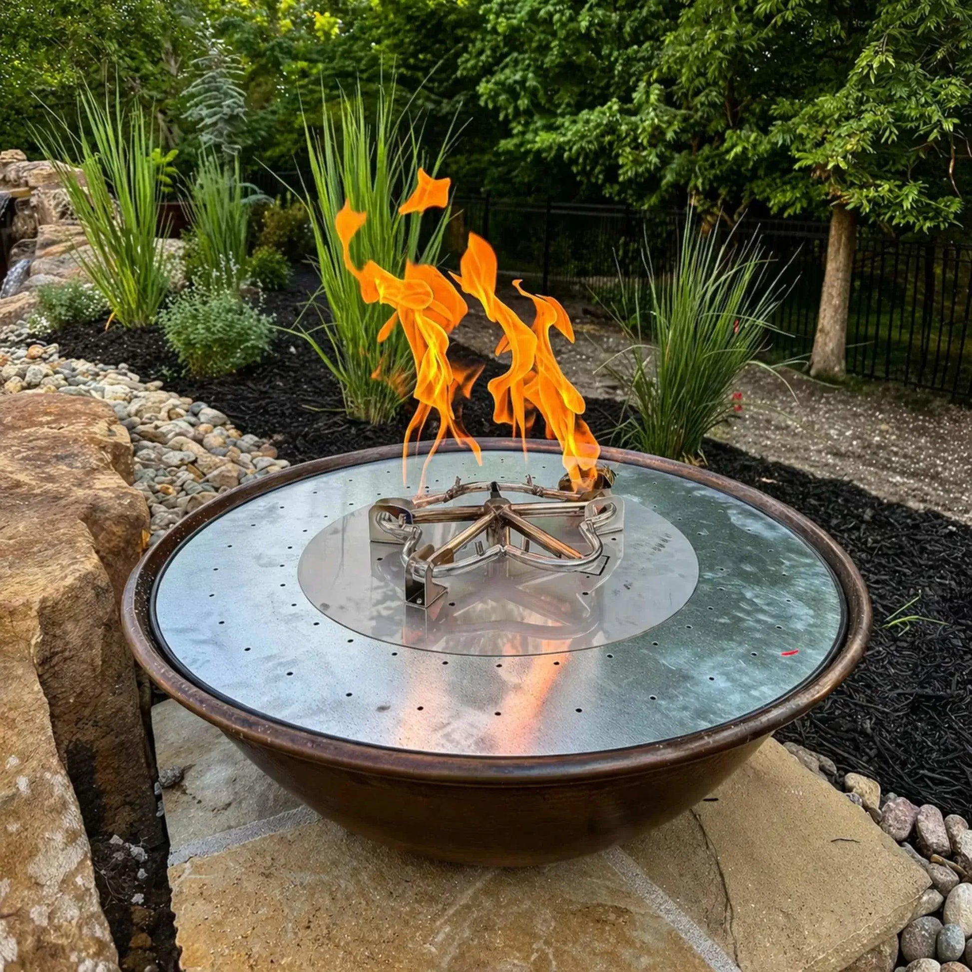 Decorative metal fire bowl with a stainless steel burner plate and visible gas burner assembly, set within a landscaped garden bed featuring ornamental grasses, rocks, and a natural stone water feature.