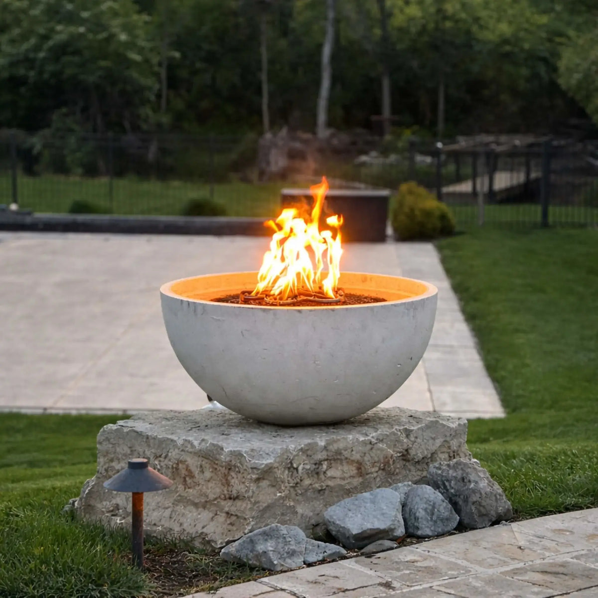 Round concrete fire bowl mounted on a stone pedestal with visible flames, placed in a manicured backyard with trimmed grass, stone accents, and a paved patio area, highlighting a clean and modern outdoor fire feature design.
