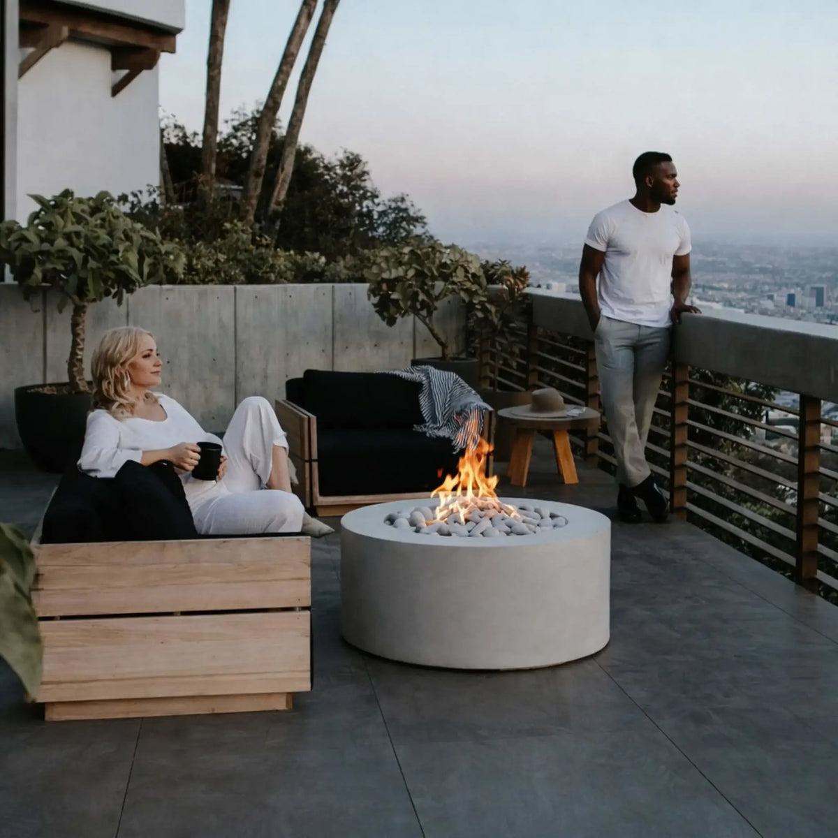 Contemporary rooftop patio with a circular gas fire pit table featuring white decorative stones and a central flame, where two people relax on outdoor furniture overlooking a city skyline at sunset.
