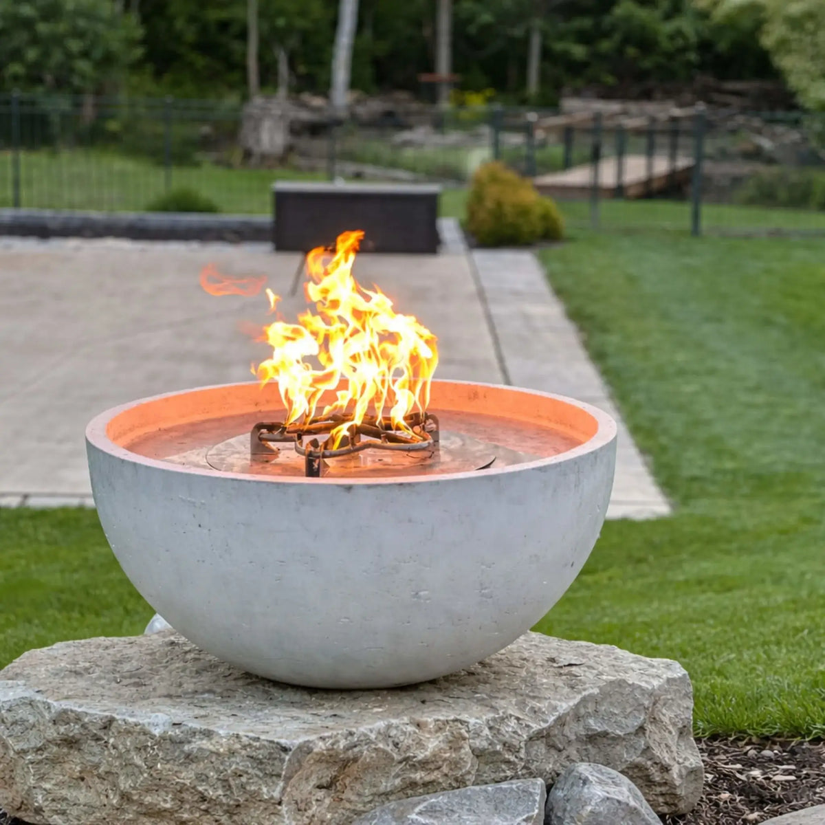 Round concrete fire bowl with a built-in penta-style gas burner producing bright flames, mounted on a natural stone pedestal in a landscaped backyard with trimmed grass, patio paving, and soft greenery in the background.