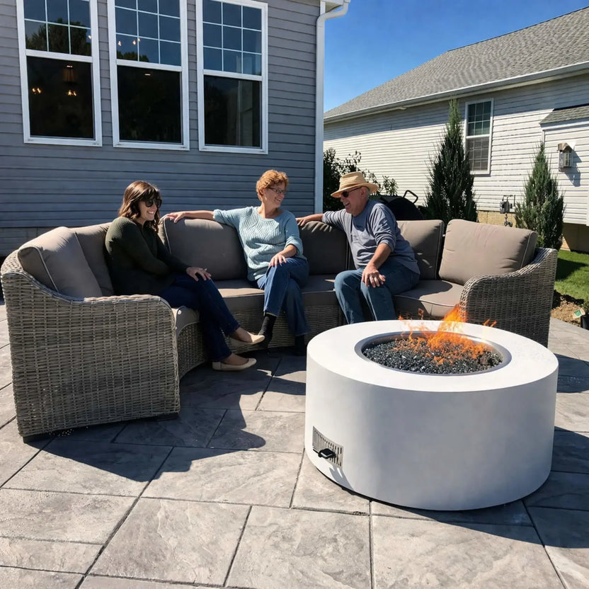 Three adults relaxing on a wicker outdoor sectional around a modern round concrete gas fire pit filled with black fire glass, set on a stamped patio beside a gray house under clear blue skies, showcasing a casual backyard gathering space.