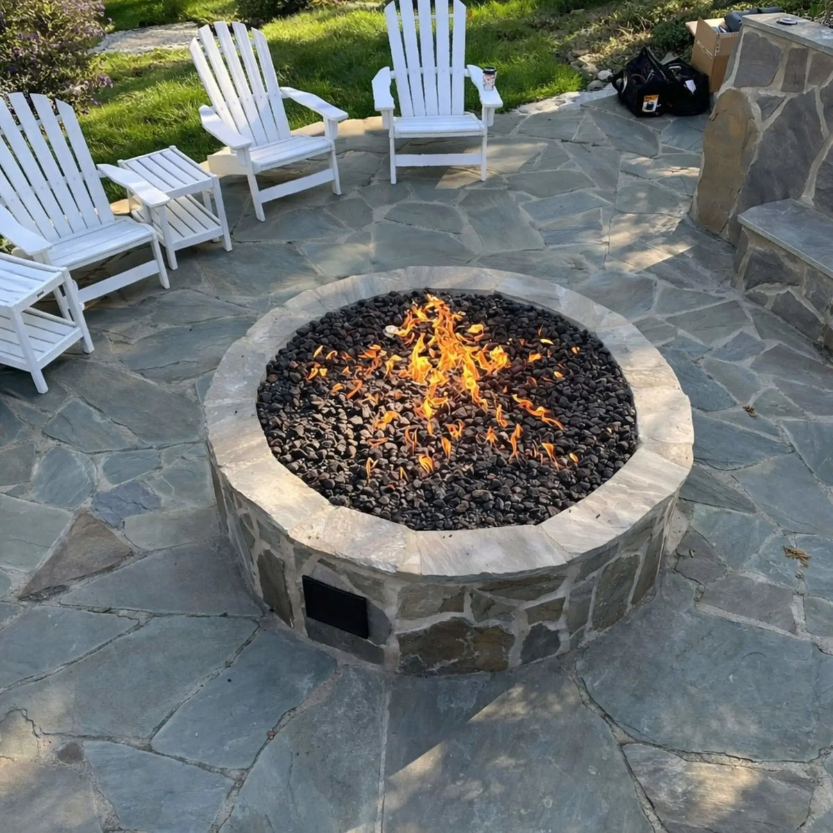 Circular stone fire pit filled with black lava rock and active flames, centered on a natural flagstone patio with white Adirondack chairs arranged around it, creating a welcoming outdoor seating area for evening gatherings.