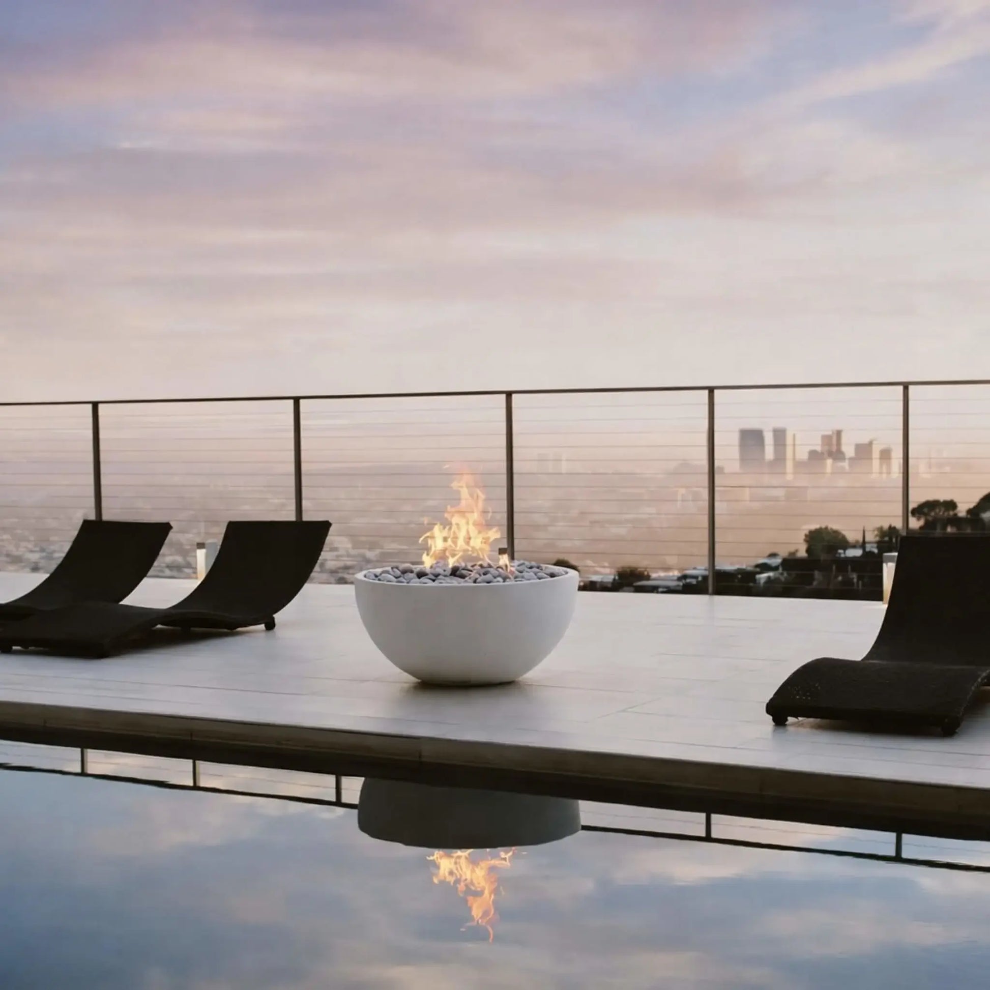 Minimalist outdoor lounge area with a round fire bowl centered on a sleek deck beside a reflective pool, featuring low-profile black loungers and a distant city skyline view at dusk, highlighting modern fire pit design.