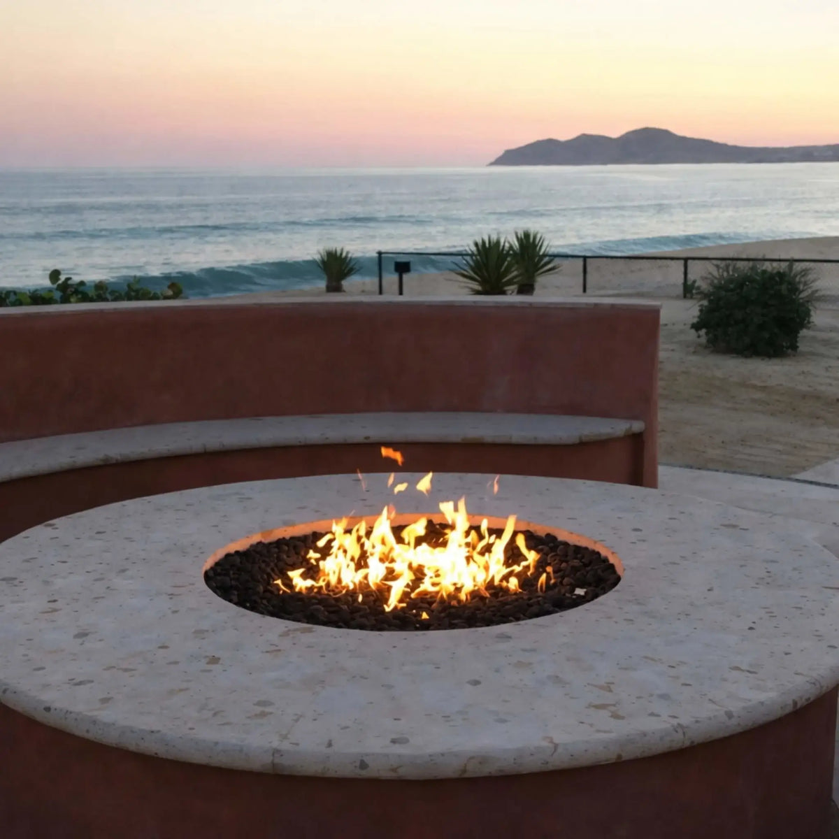 Beachfront fire pit built into a curved stucco seating wall, with a circular burner producing bright flames over lava rock, set against a scenic ocean backdrop at sunset with sandy terrain and coastal vegetation.
