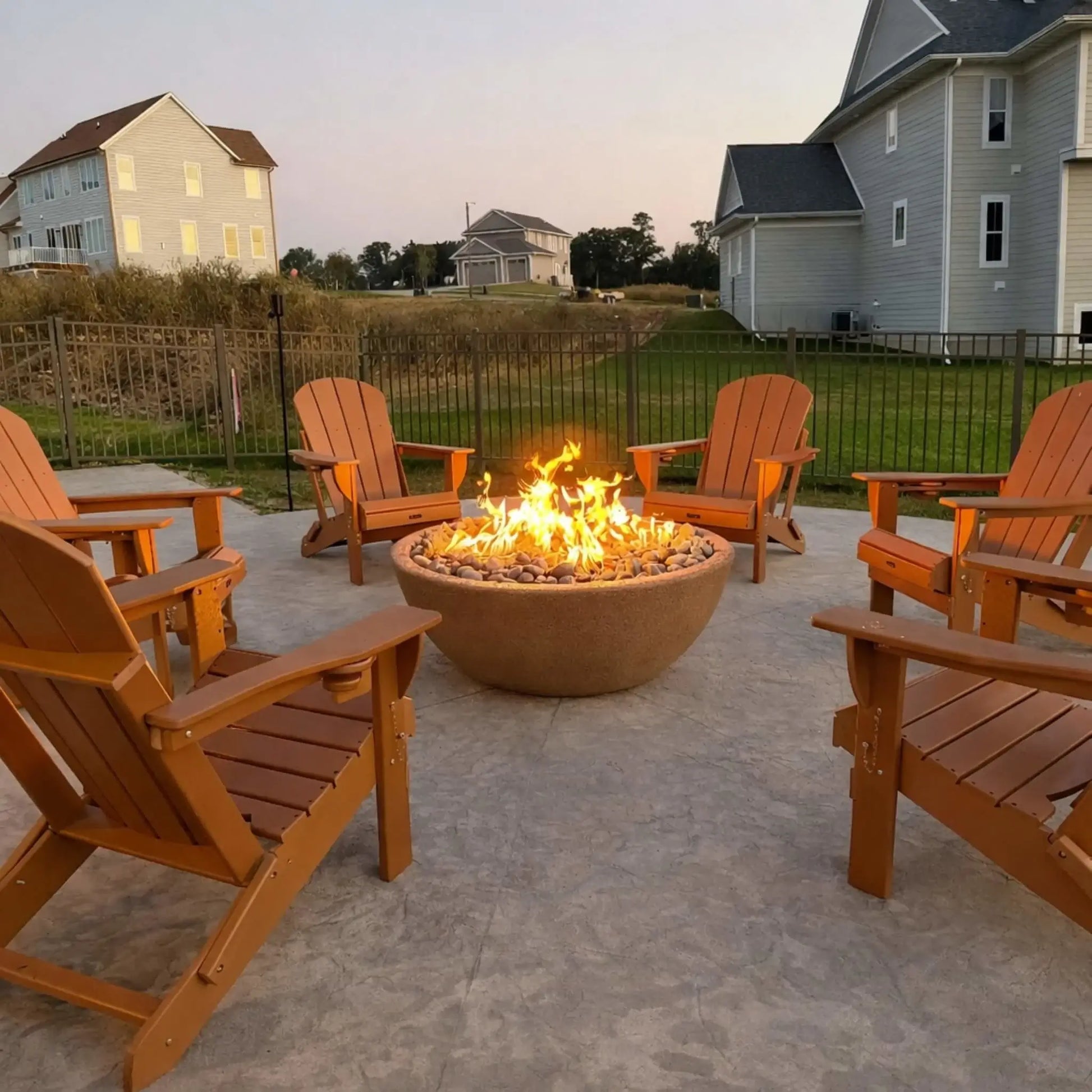 Suburban backyard patio featuring a round concrete fire pit with a strong gas flame surrounded by multiple wooden Adirondack chairs, positioned on a stamped concrete surface with nearby homes and fenced yard.