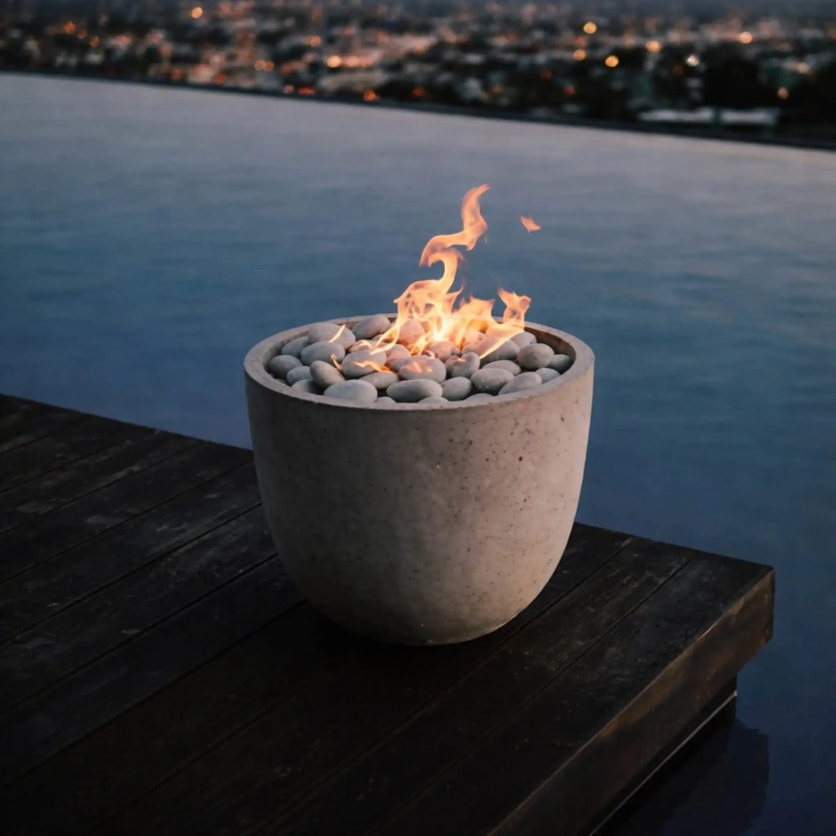Compact round concrete fire bowl with smooth river stones and a clean-burning flame placed on a wooden deck overlooking a calm infinity pool and distant city lights, creating a serene modern outdoor ambiance.
