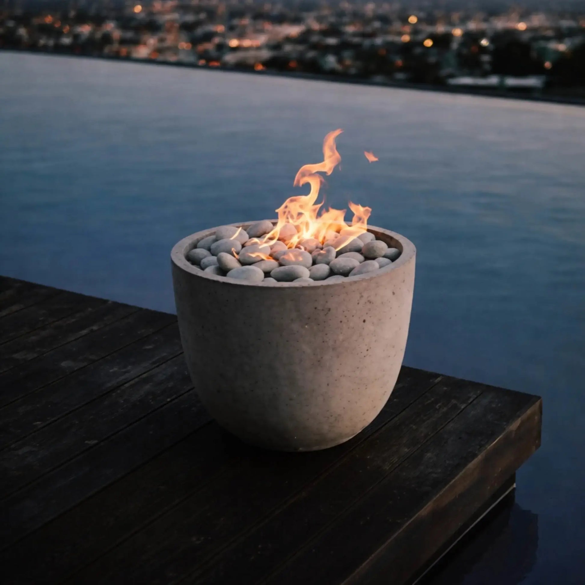 Compact round concrete fire bowl with smooth river stones and a clean-burning flame placed on a wooden deck overlooking a calm infinity pool and distant city lights, creating a serene modern outdoor ambiance.