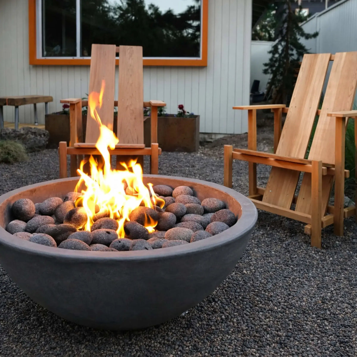 Modern backyard fire feature with a wide concrete fire bowl filled with natural stones and active flames, surrounded by wooden Adirondack chairs on gravel landscaping near a contemporary home exterior.