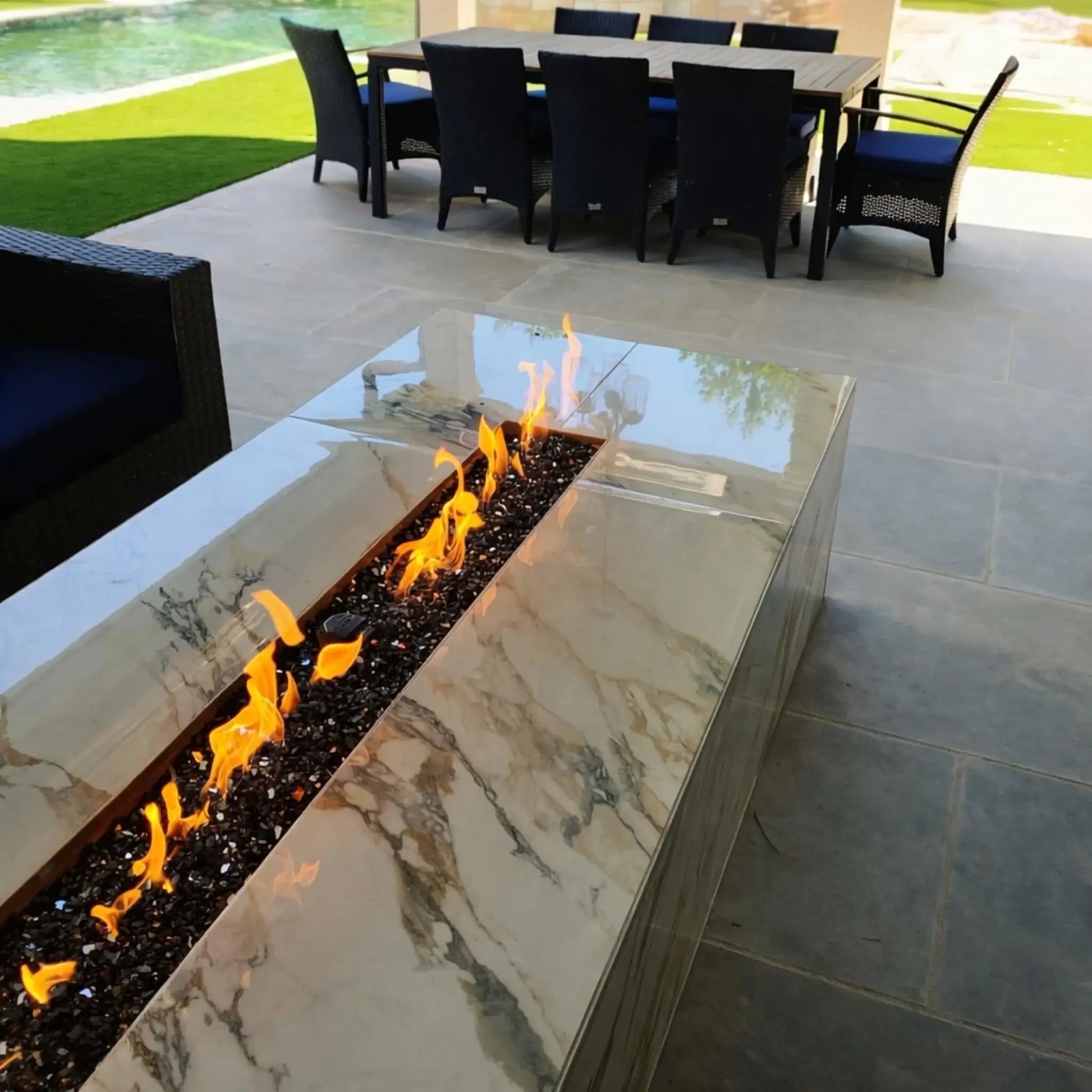 Modern rectangular outdoor fire pit table with a polished marble-style finish featuring a long linear burner filled with black tempered fire glass, producing vibrant yellow flames on a covered patio with dining furniture overlooking a pool and landscaped backyard.
