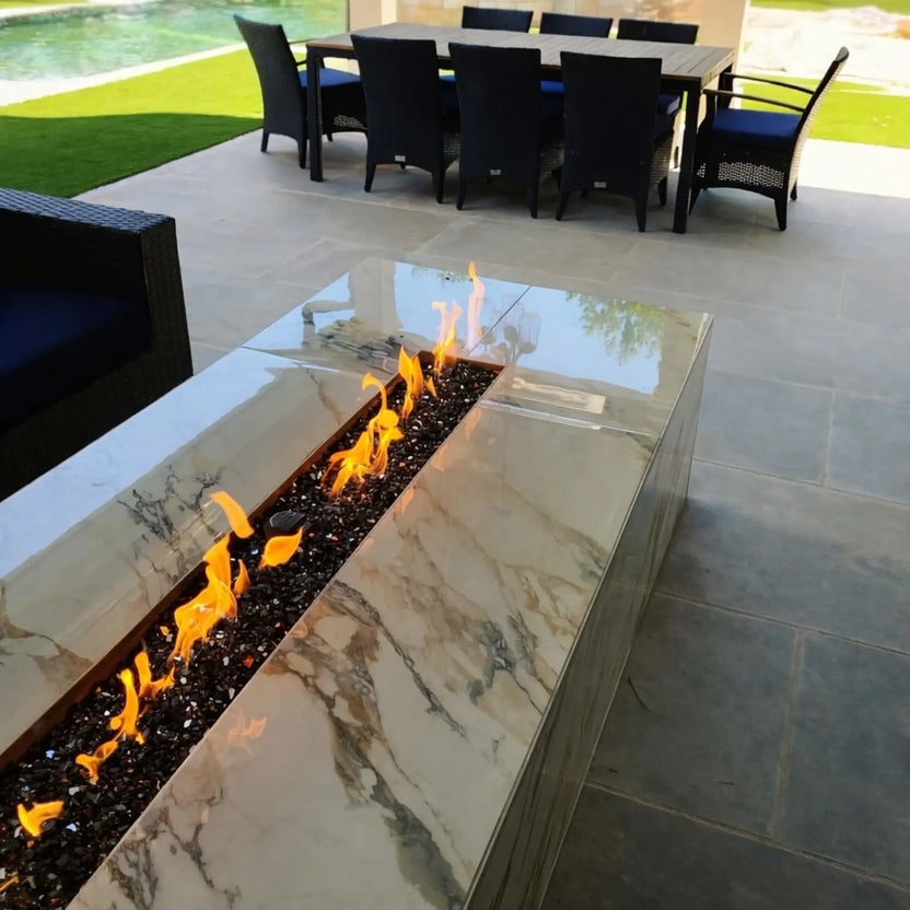 Modern rectangular outdoor fire pit table with a polished marble-style finish featuring a long linear burner filled with black tempered fire glass, producing vibrant yellow flames on a covered patio with dining furniture overlooking a pool and landscaped backyard.