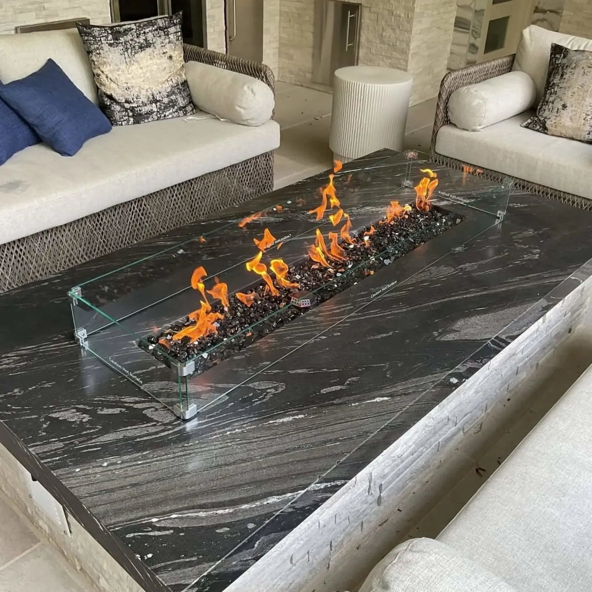 Luxury outdoor seating area with a large rectangular fire pit table finished in dark stone, showcasing a linear burner filled with black fire glass and surrounded by cushioned wicker sofas, accented by a clear glass wind guard and contemporary patio design.